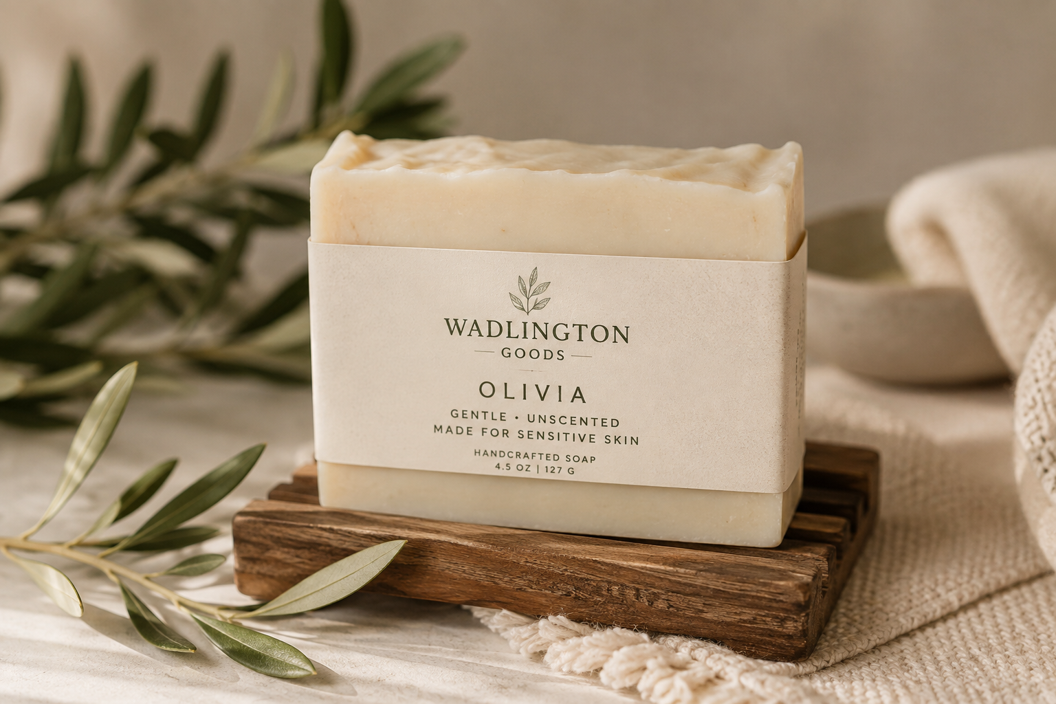 A bar of handmade soap labeled "Olivia" from Wadlington Goods, resting on a wooden platform with green foliage and a cream-colored cloth in the background.