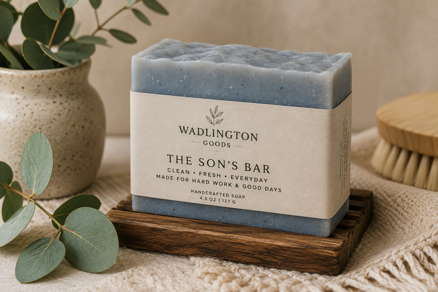 A bar of handcrafted soap labeled "Wadlington Goods, The Son's Bar, Handmade Soap, 4.5 oz" placed on a small wooden platform with a tea towel. There is a potted plant and a wooden scrub brush nearby.