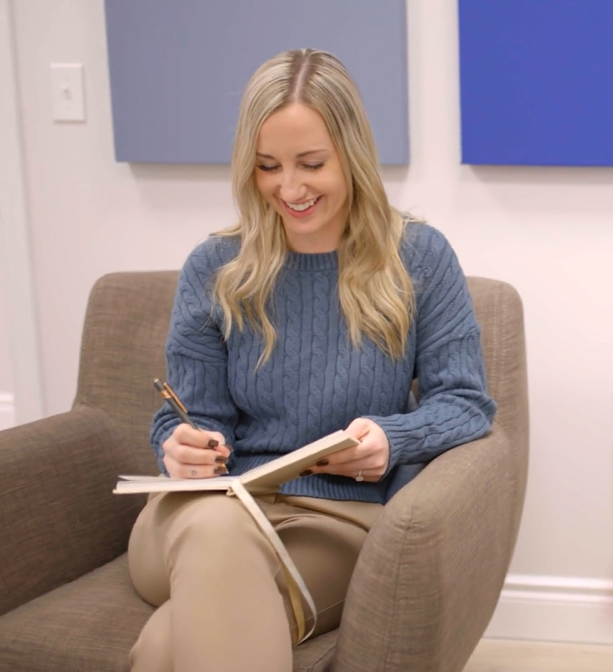 A woman with long blonde hair sitting on a beige chair, smiling and writing in a notebook.