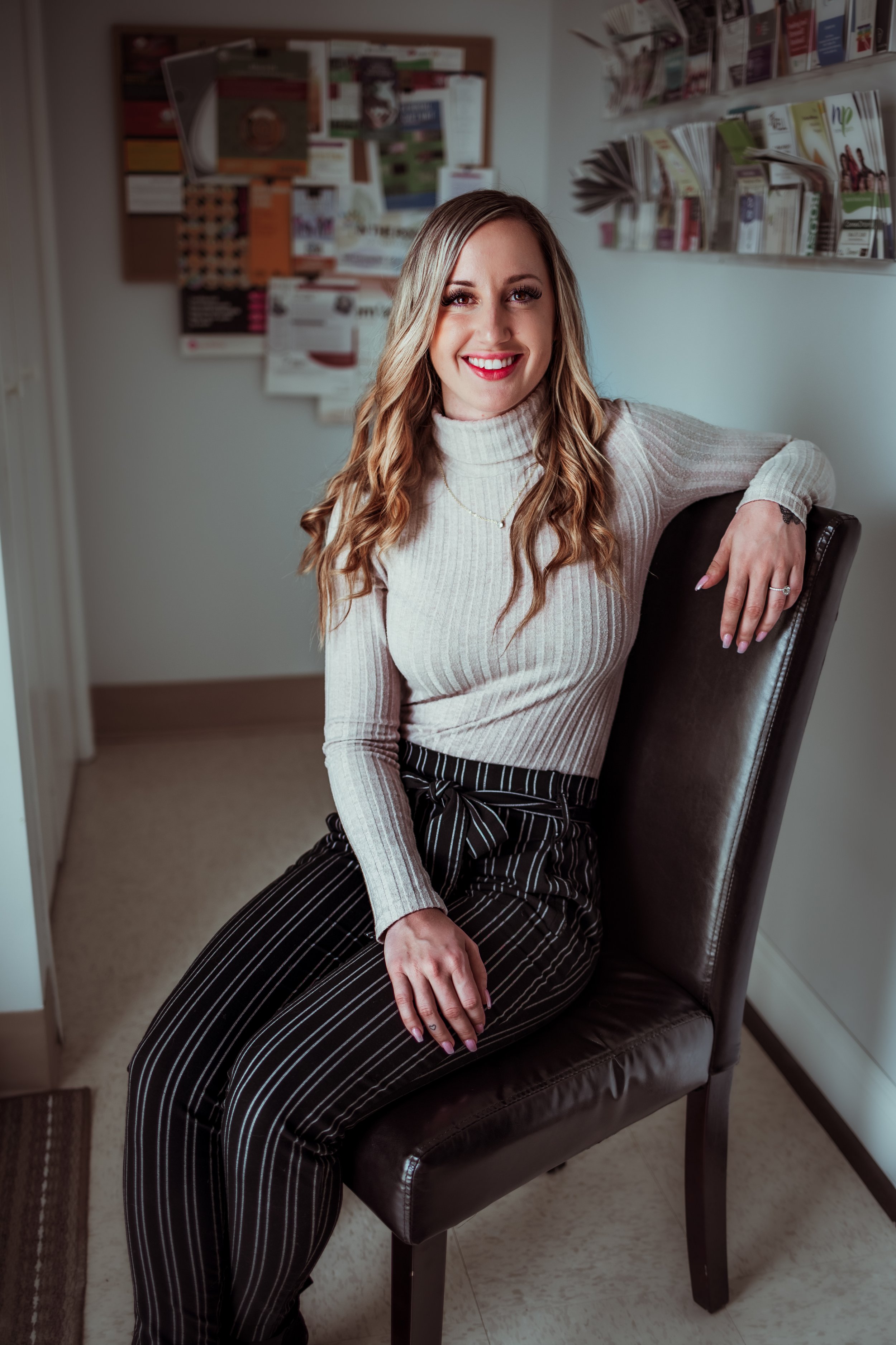A woman with long wavy hair, wearing a light-colored turtleneck sweater and black pants with white pinstripes, sitting on a dark brown chair in an indoor setting with bulletin boards and magazines or pamphlets on the wall behind her, smiling at the camera.