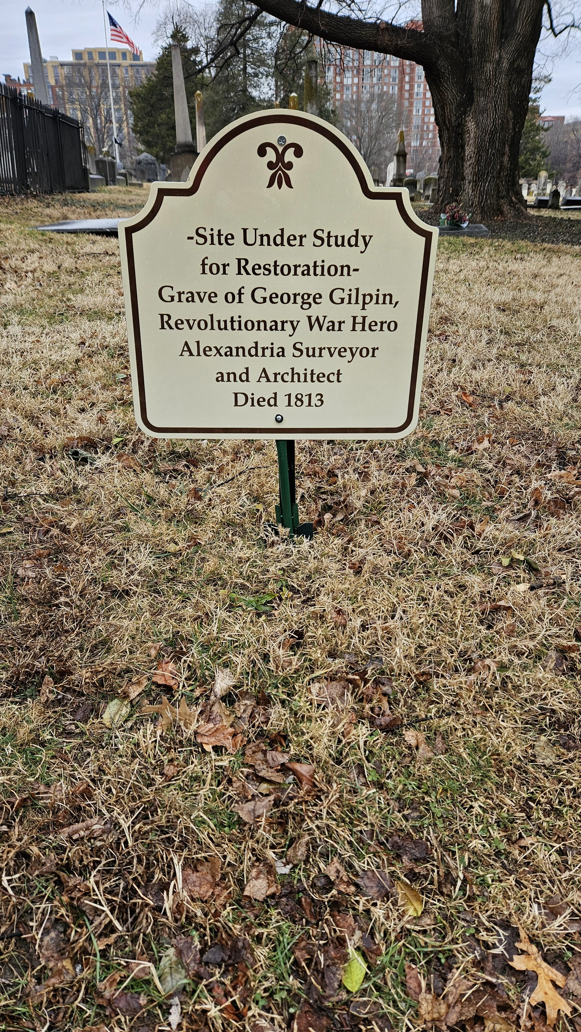 A graveyard with a sign indicating a site under study for restoration. The sign states it is the grave of George Gilpin, a Revolutionary War hero, surveyor, and architect, who died in 1813. There are trees, grass, gravestones, and a large building with an American flag in the background.