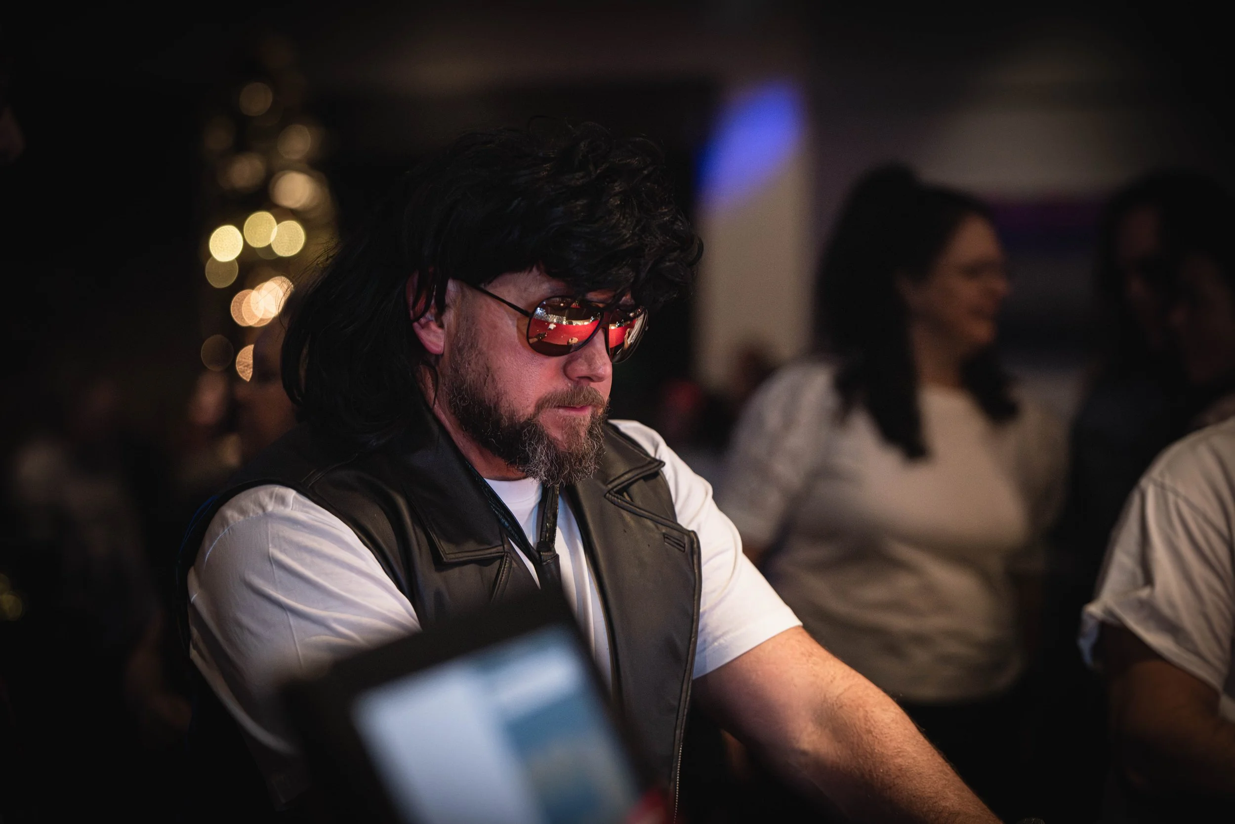 Homme avec lunettes de soleil réfléchissant, cheveux noirs, barbe, portant une veste en cuir blanc, assis à une table dans un environnement sombre avec d'autres personnes en arrière-plan.