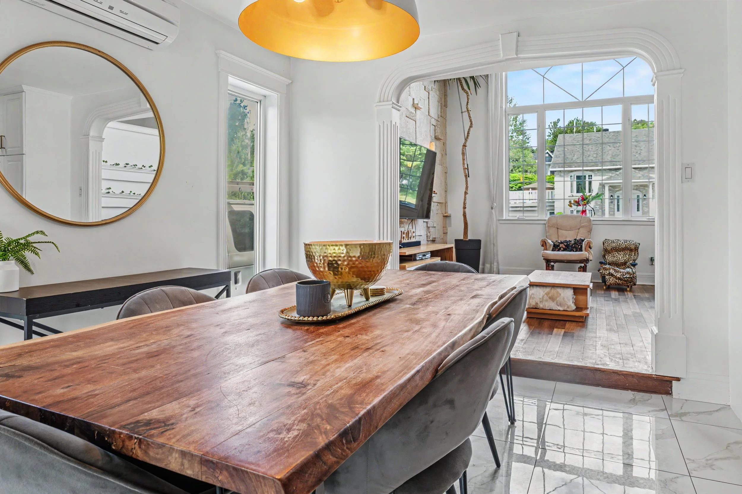 Une salle à manger lumineuse avec un grand meuble en bois, un miroir rond encadré de doré, des fenêtres laissant entrer la lumière naturelle, et des décorations comme un chandelier doré, une plante, et des chaises en velours autour d'une table en bois.