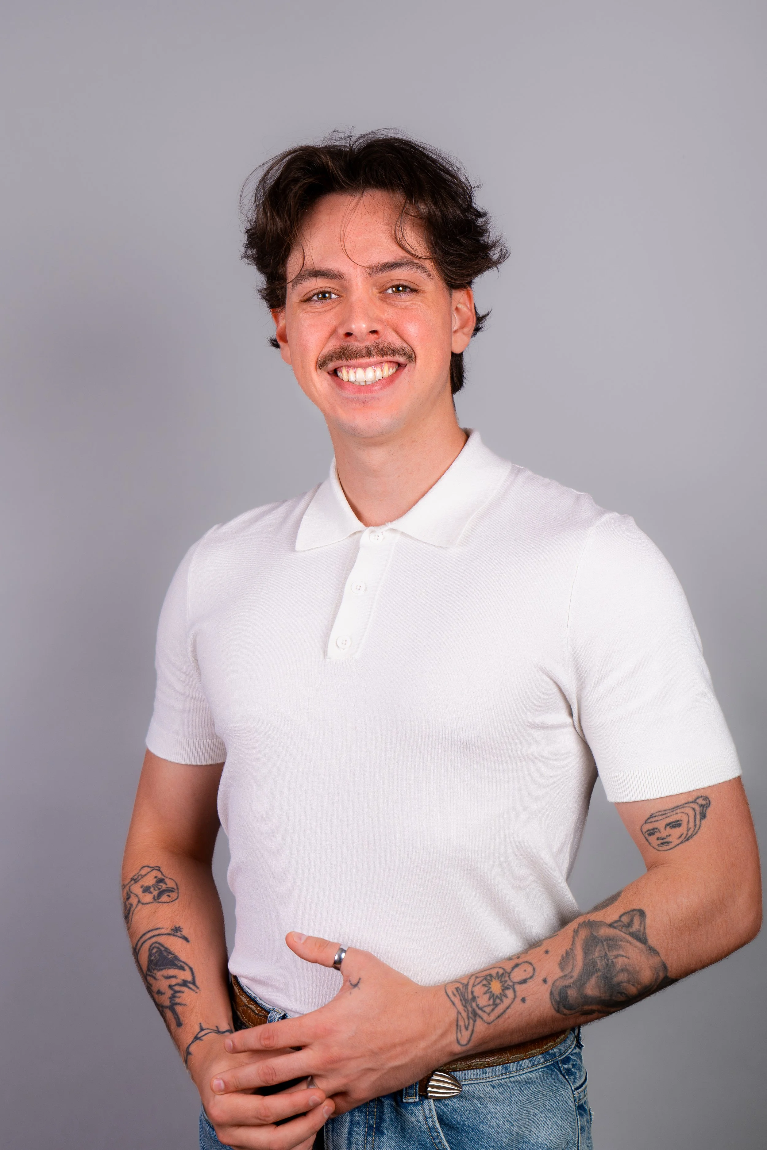 Portrait d'un homme souriant avec des tatouages sur les bras, portant un polo blanc, contre un fond gris.