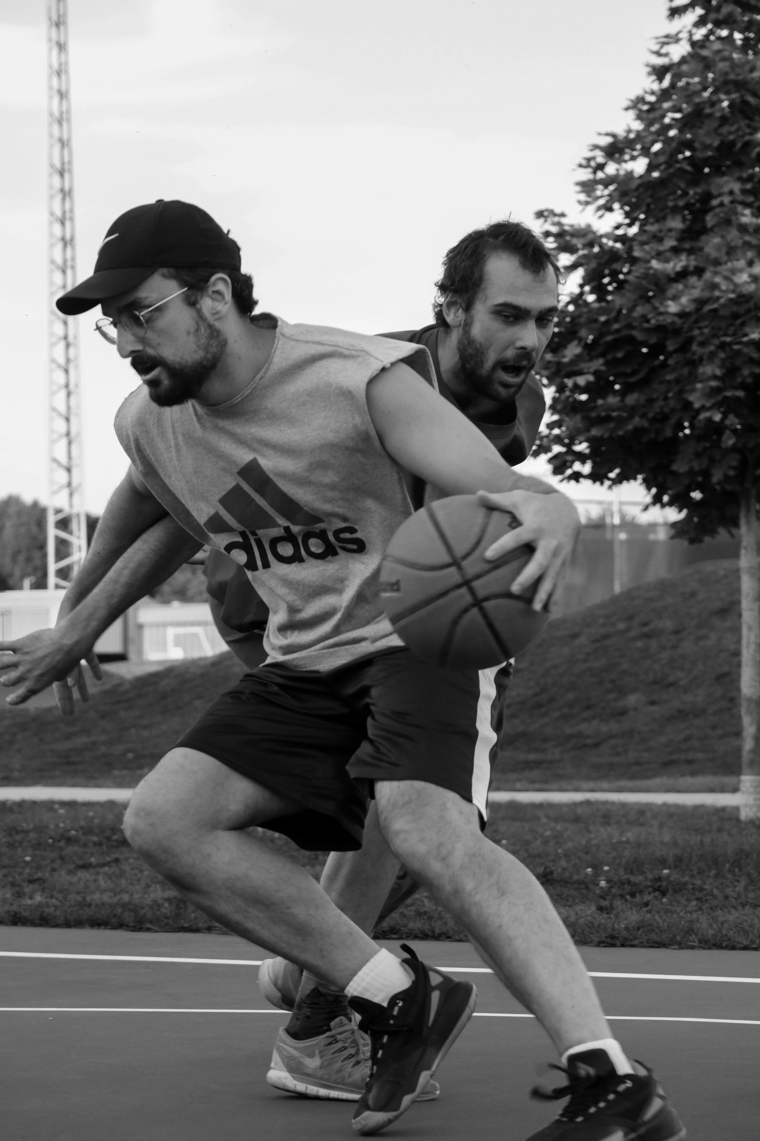 Deux hommes jouent au basketball sur un terrain en extérieur, en mouvement. La scène est capturée en noir et blanc.