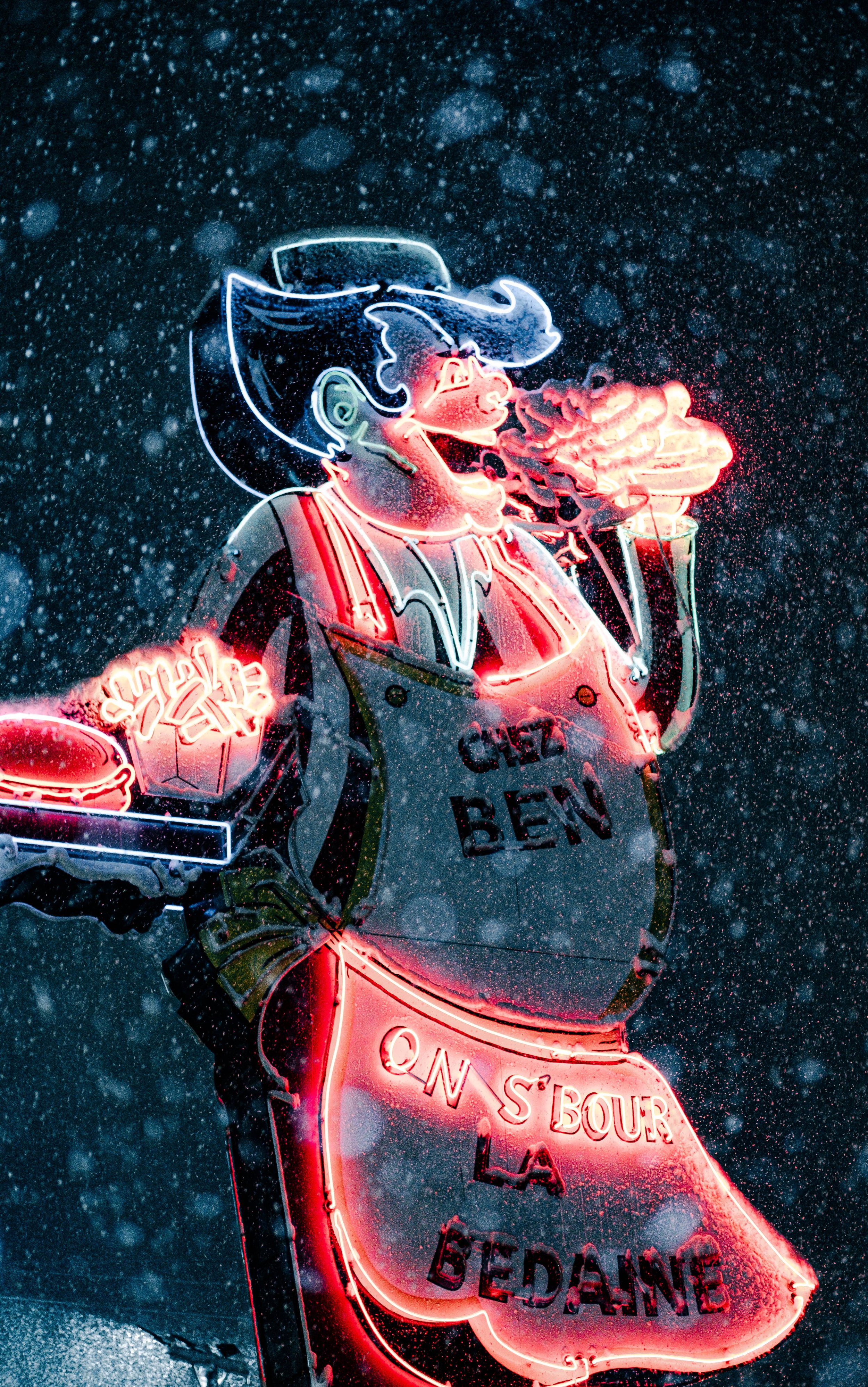Sign neon représentant une femme dans un style cartoon, fumant une cigarette, sous la pluie, avec des phrases en français comme « QUEZ' BEN » et « ON S'BOUR »