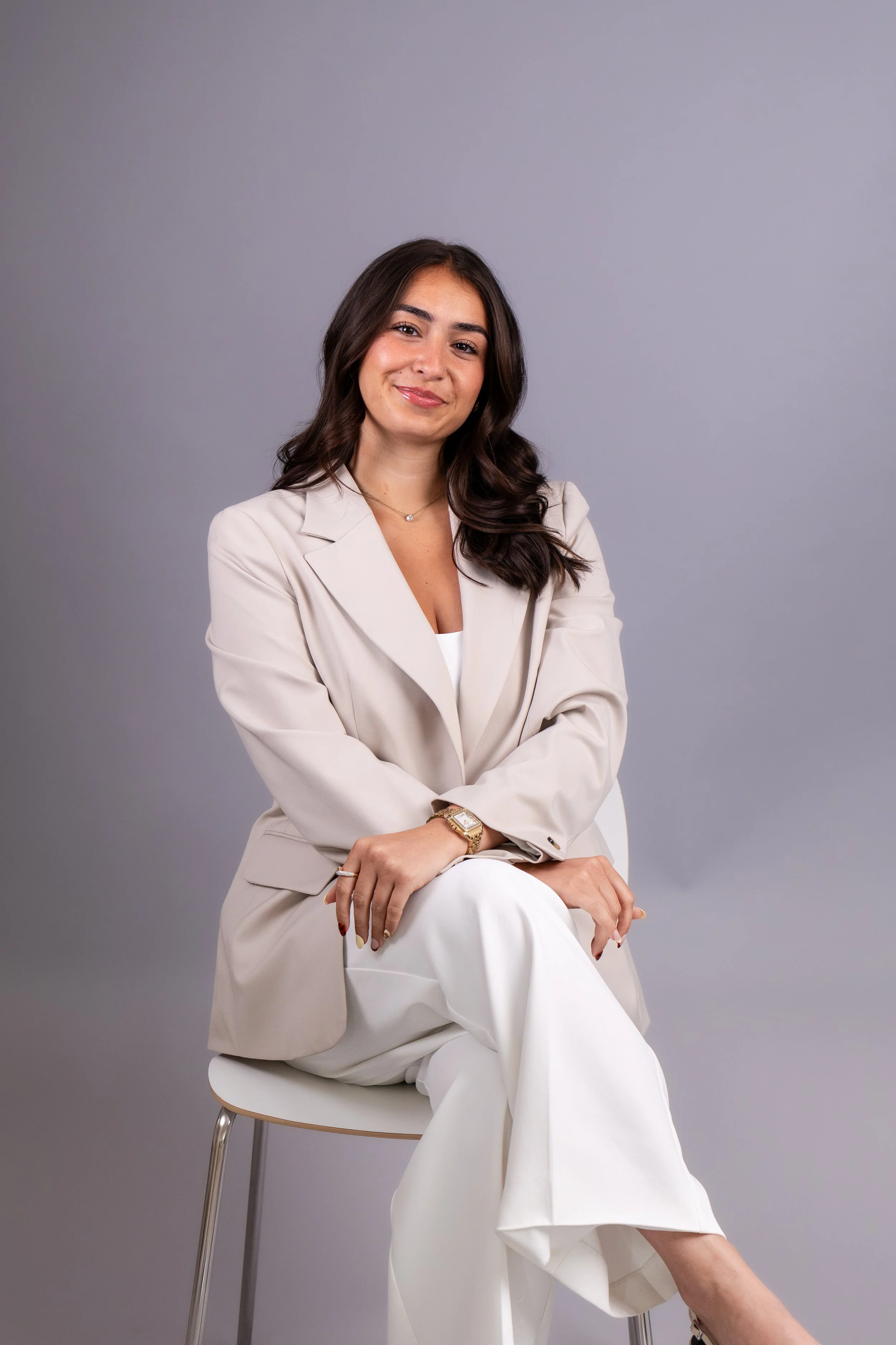 Une femme assise sur une chaise en bois avec un fond gris clair, portant un tailleur beige et des vêtements blancs, souriant et croisant les bras.