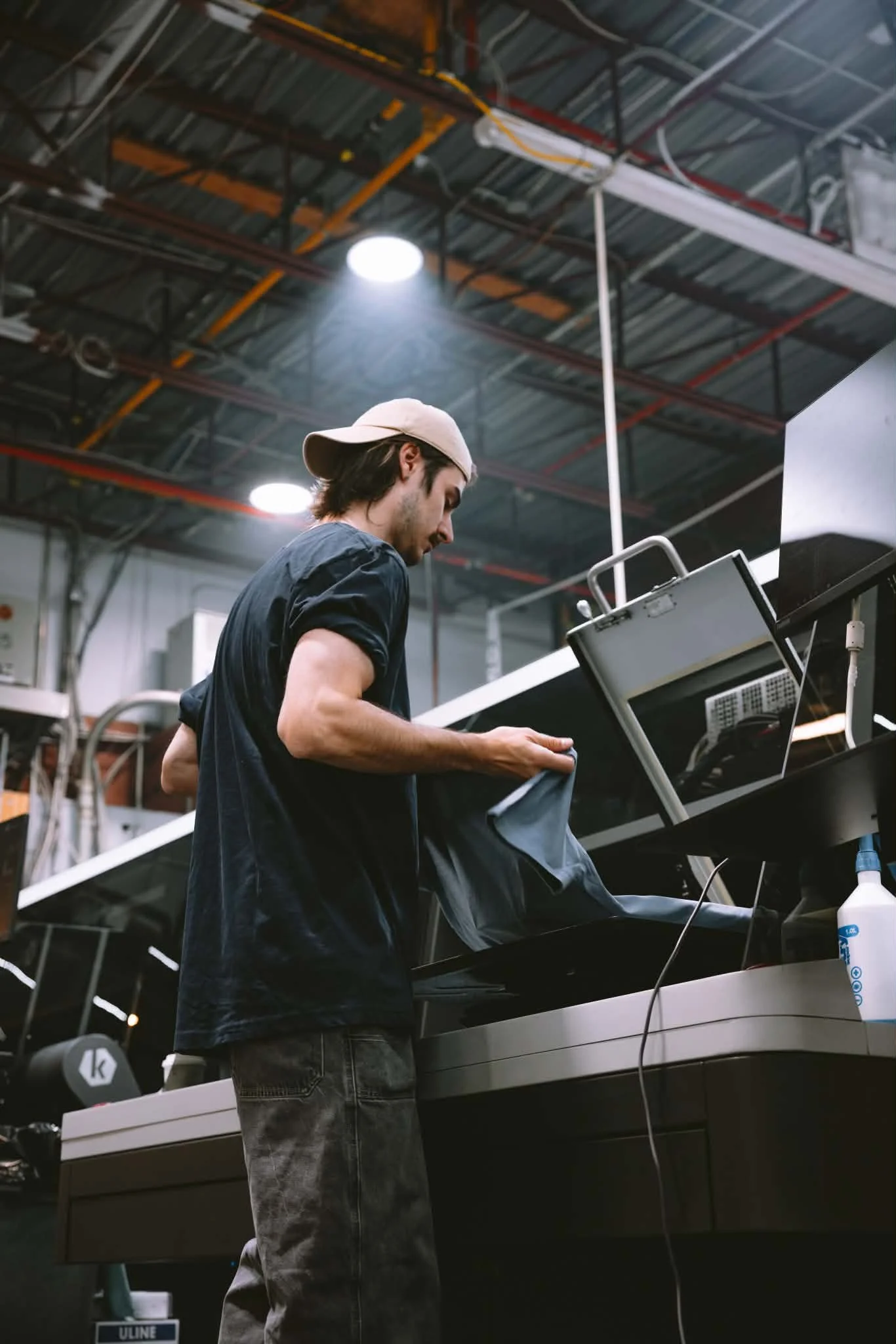 Un jeune homme travaille sur un piano dans un atelier industriel, portant une casquette beige et un t-shirt noir, avec des luminaires suspendus au plafond.