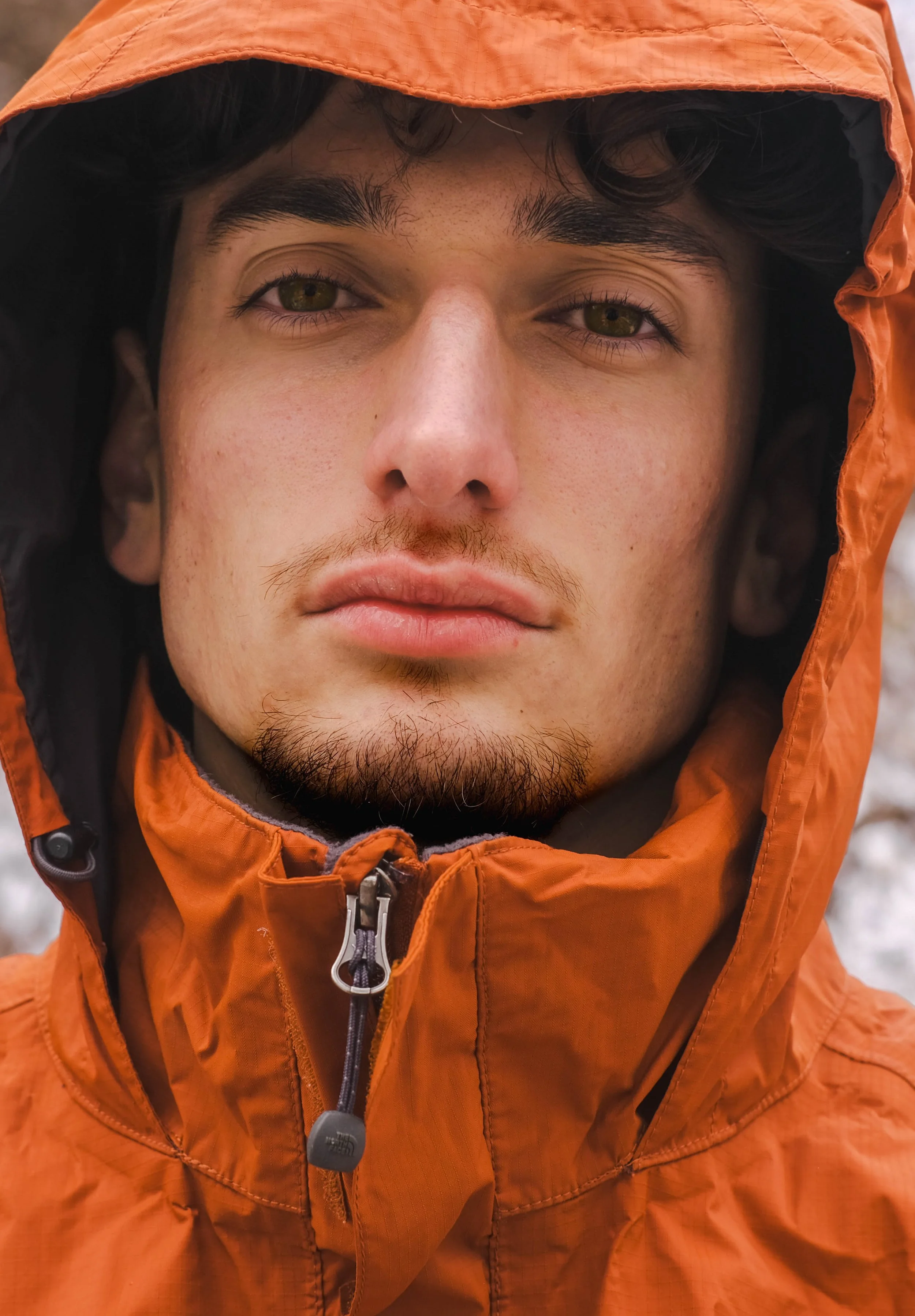 Un homme avec des cheveux noirs, barbe légère, portant une veste orange avec capuche, dans un environnement extérieur.
