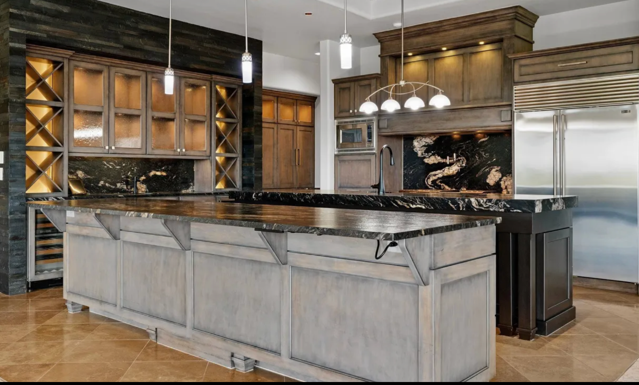 Lux360 Clean's cleaning of a modern kitchen with wooden cabinets, a large black marble island with a wood countertop, built-in wine racks, stainless steel refrigerator, and black backsplash with a marble design. Pendant lights hang above the island.