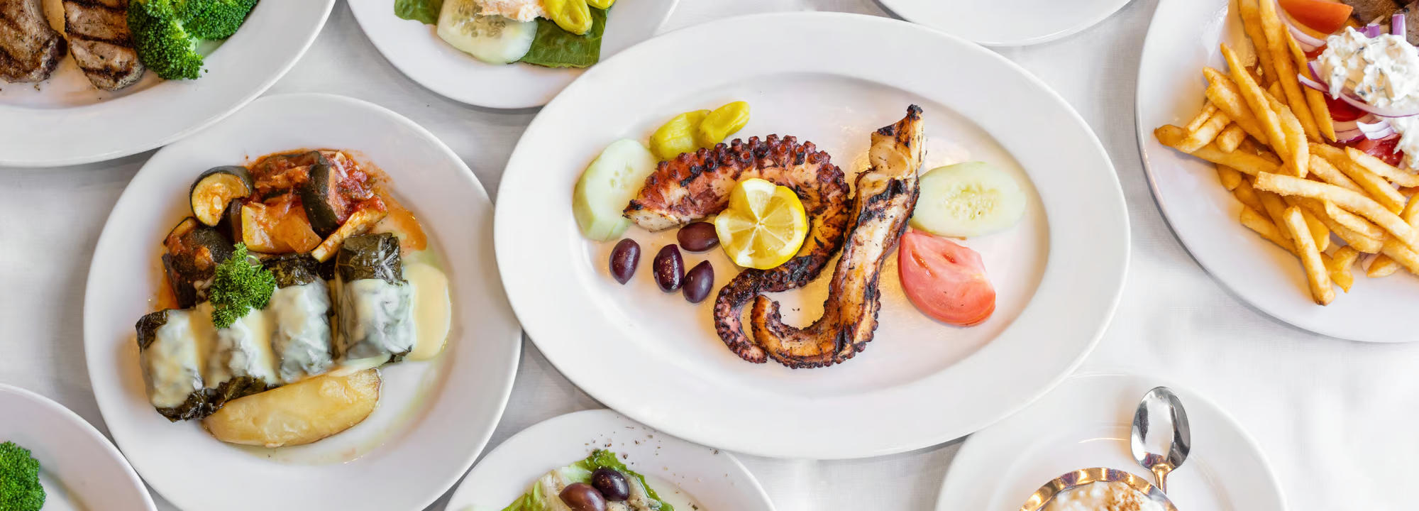 Various plates of food including grilled meats with vegetables, an octopus dish with lemon and olives, and a plate of French fries with toppings, on a white tablecloth.
