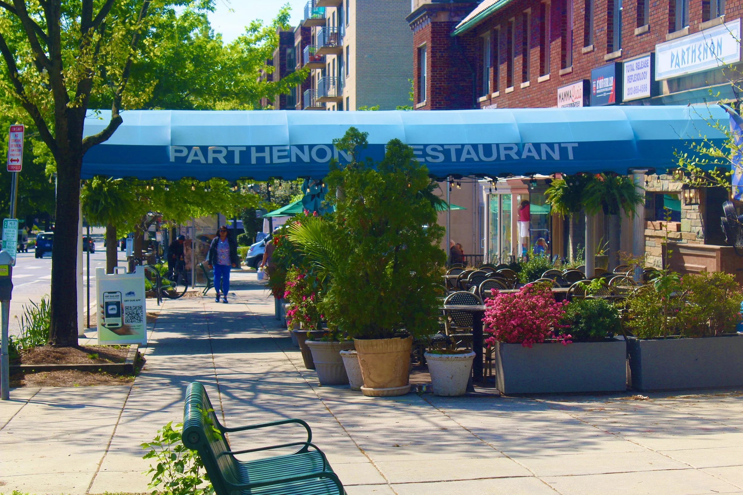 Outdoor sidewalk scene with potted plants, pink flowers, green trees, a shaded restaurant patio with chairs, and a blue awning with white text that reads "Parthenon Restaurant". There are a few people walking on the sidewalk and brick buildings in the background.
