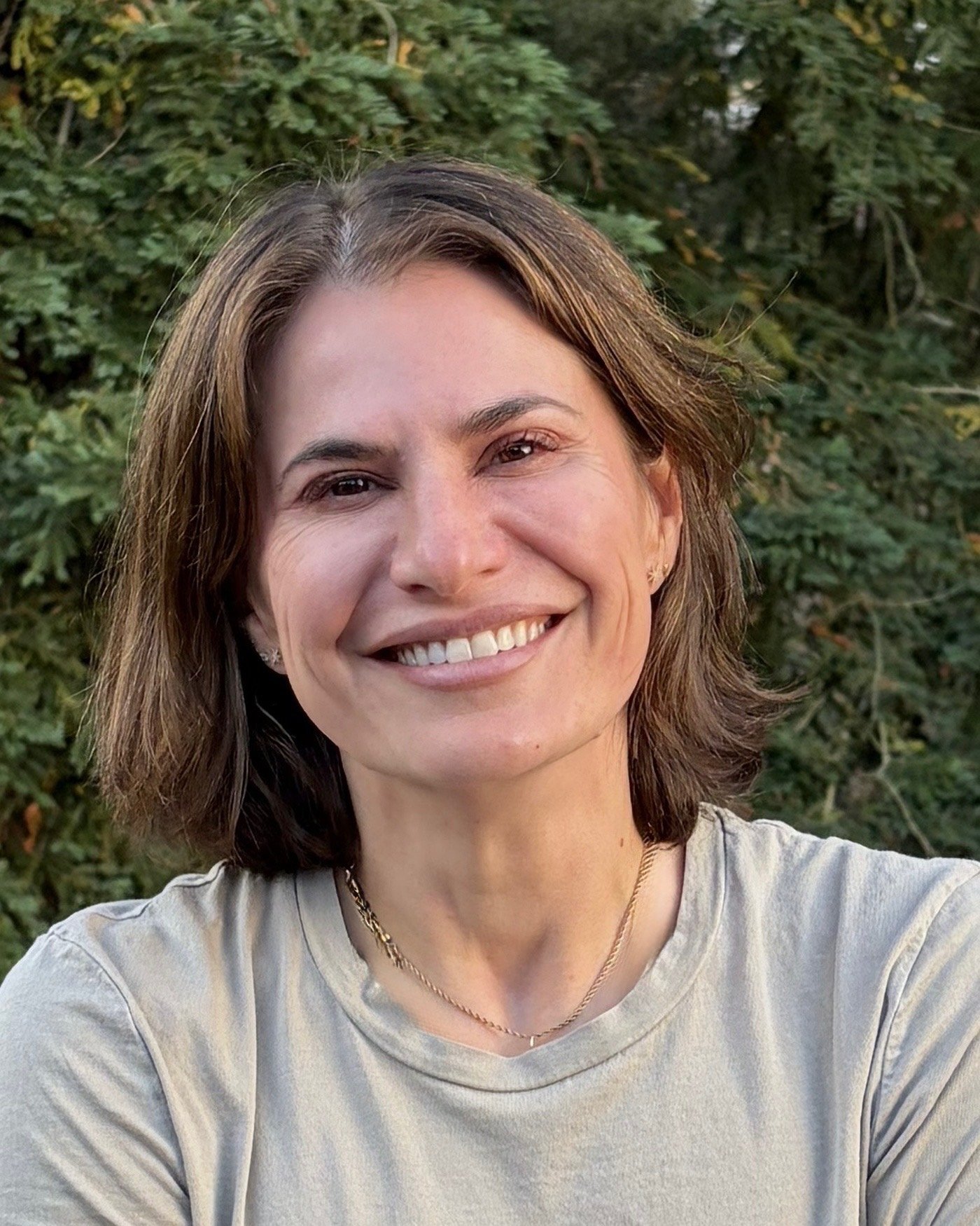 A smiling woman with brown hair taking a selfie outdoors in front of green foliage.