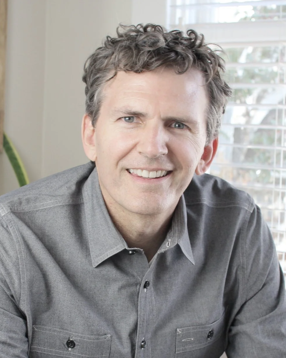 A man with curly brown hair and blue eyes smiling, wearing a gray button-up shirt, sitting indoors near a window with blinds.