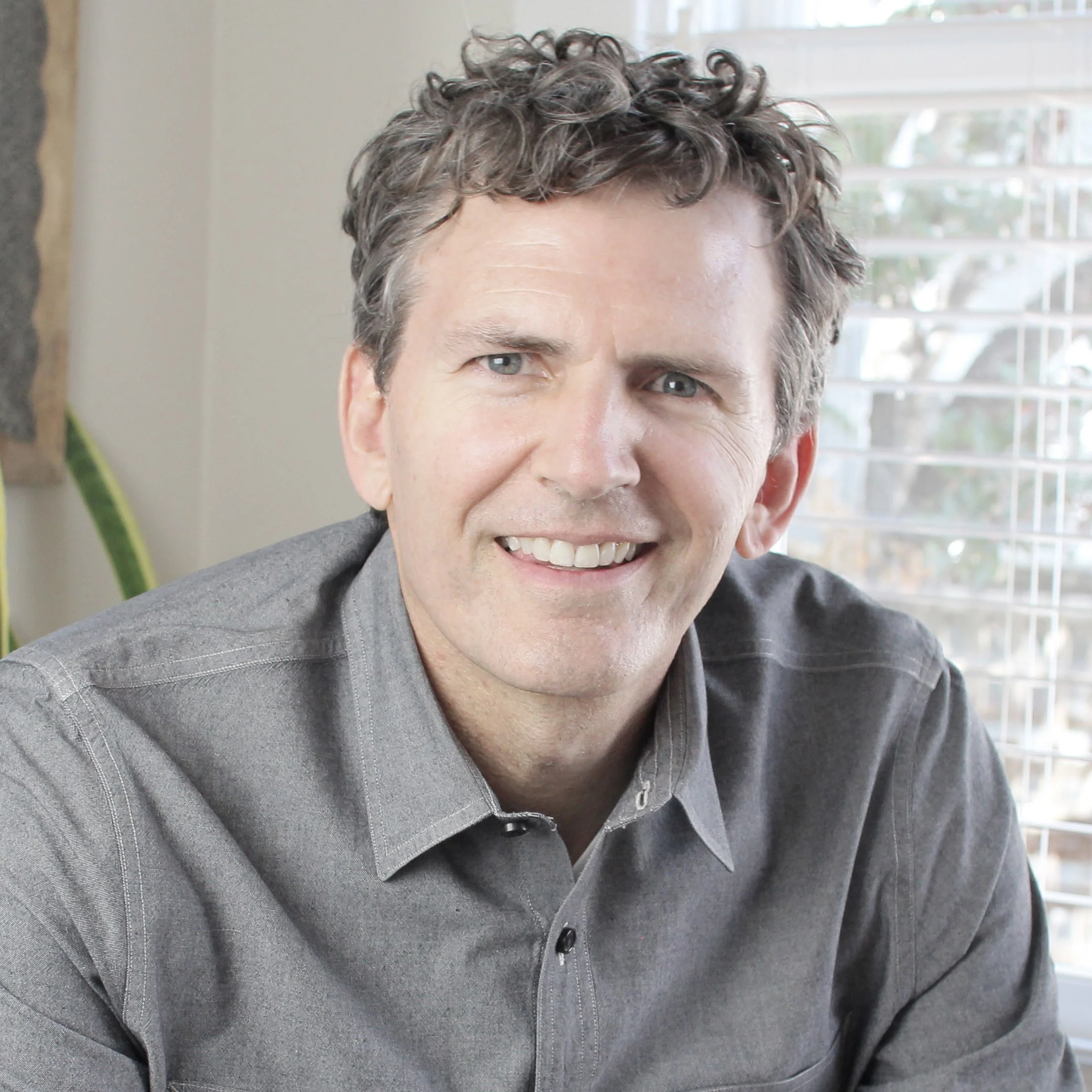Close-up of a smiling man with curly hair wearing a gray button-up shirt in front of a window with blinds.