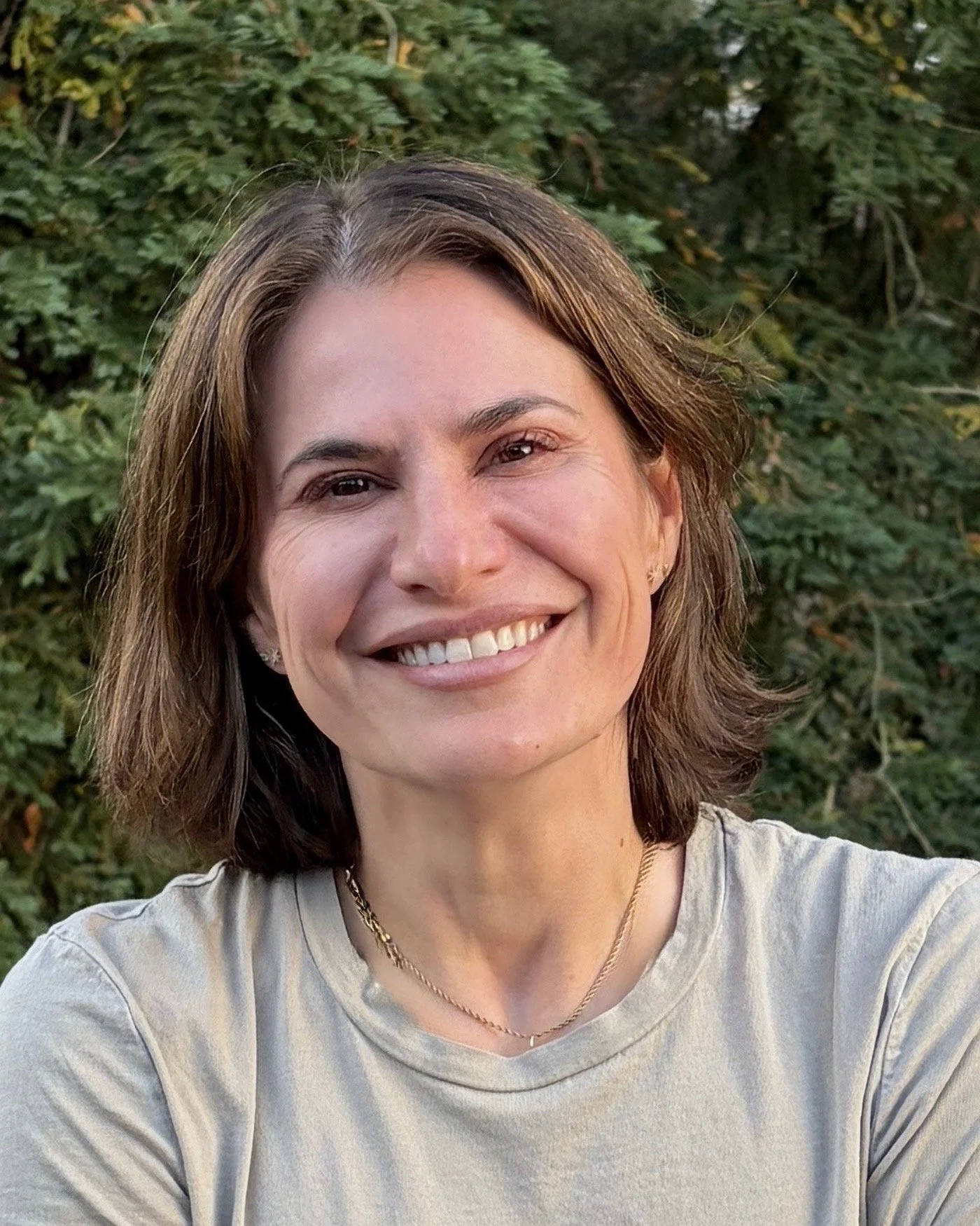 A smiling woman with short brown hair and a beige shirt outdoors with green foliage in the background.