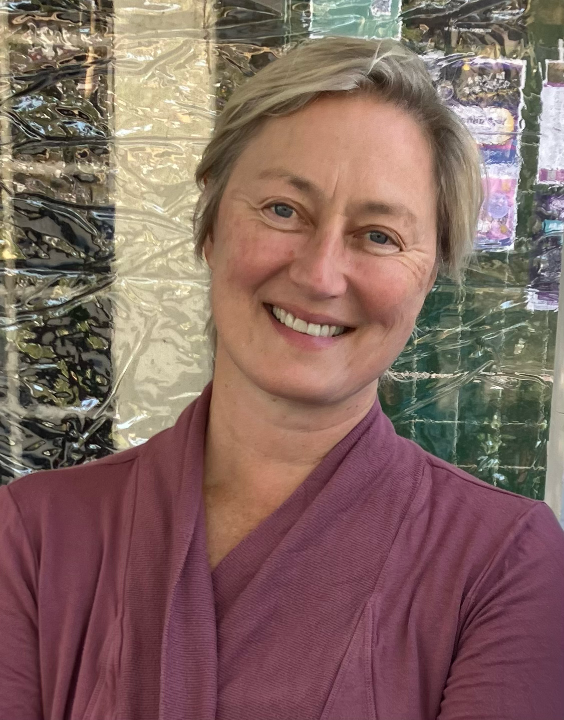 A smiling woman with short blonde hair, wearing a pink or mauve uniform, standing in front of a shiny, reflective background that appears to be wrapped in metallic or reflective material.