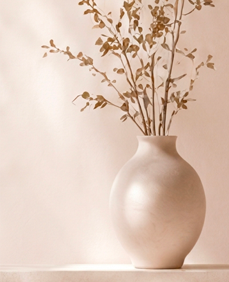 A large, plain, oval-shaped beige vase with dried branches and leaves inside, placed on a surface against a light-colored wall.