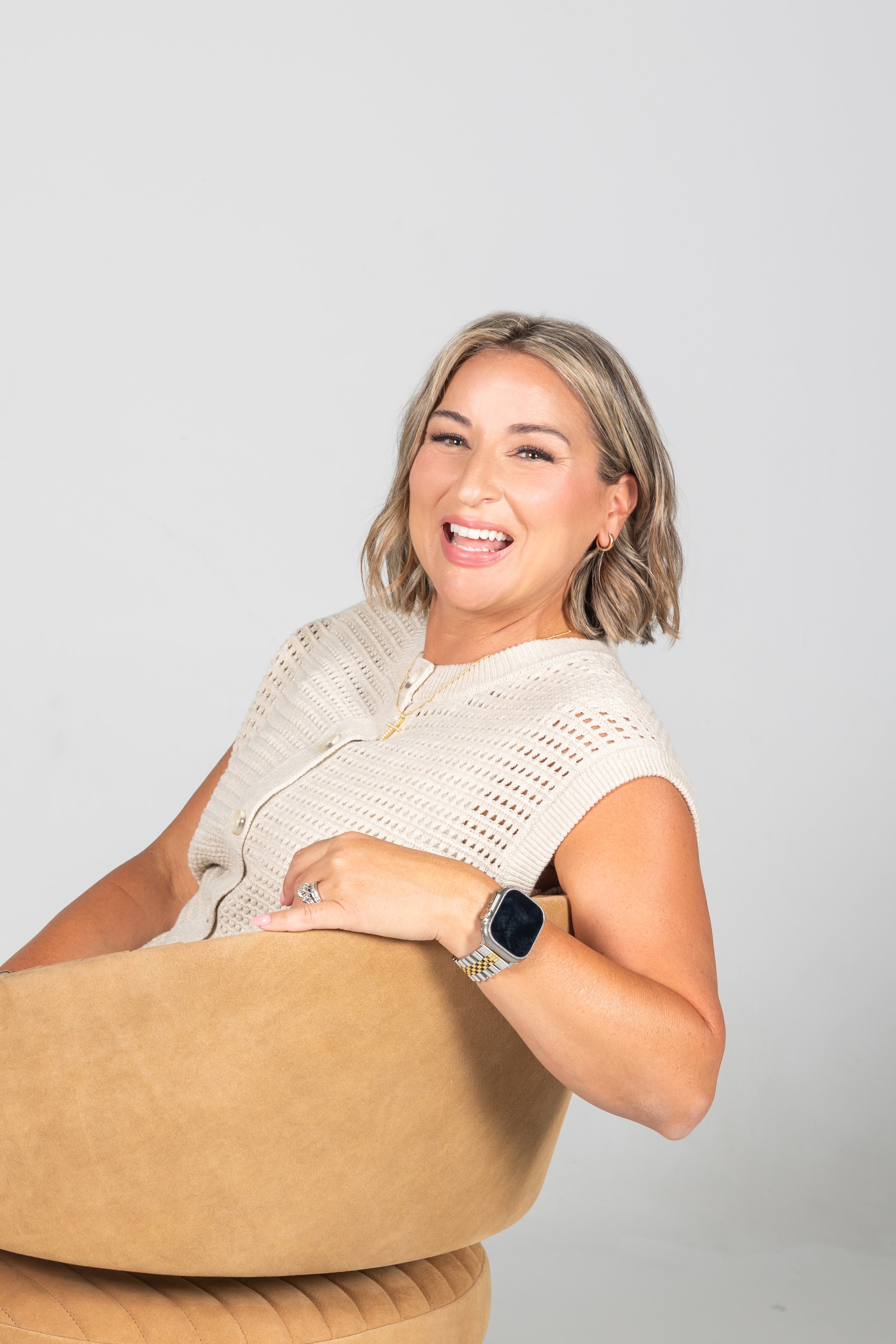A smiling woman with blonde hair wearing a beige sleeveless top, holding a large cardboard box in her arm, and sitting on a tan cushioned chair against a plain light gray background.