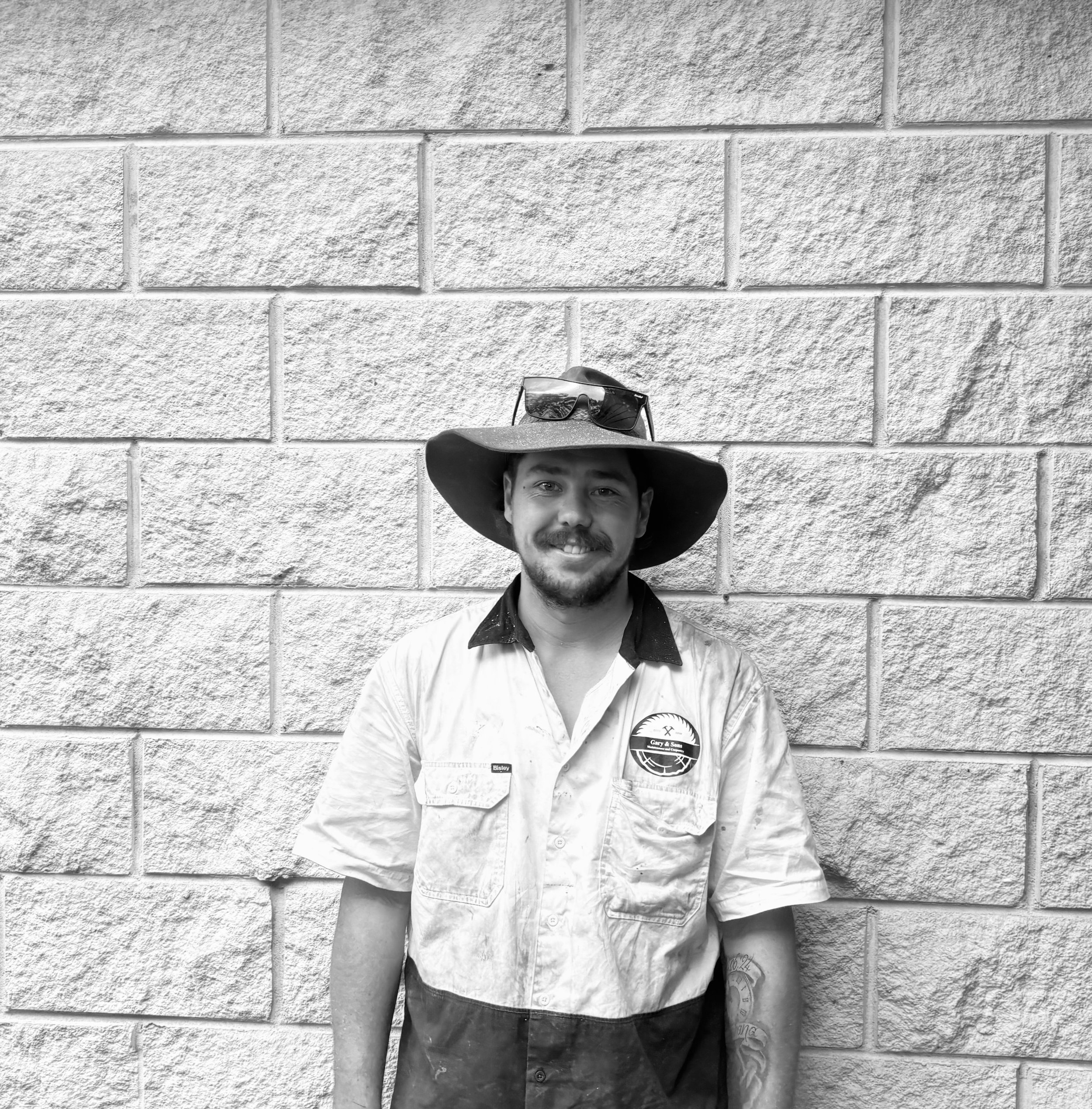 Black and white photo of a young man wearing a wide-brimmed hat, sunglasses, a scarf, and button-up shirt standing in front of a textured brick wall.