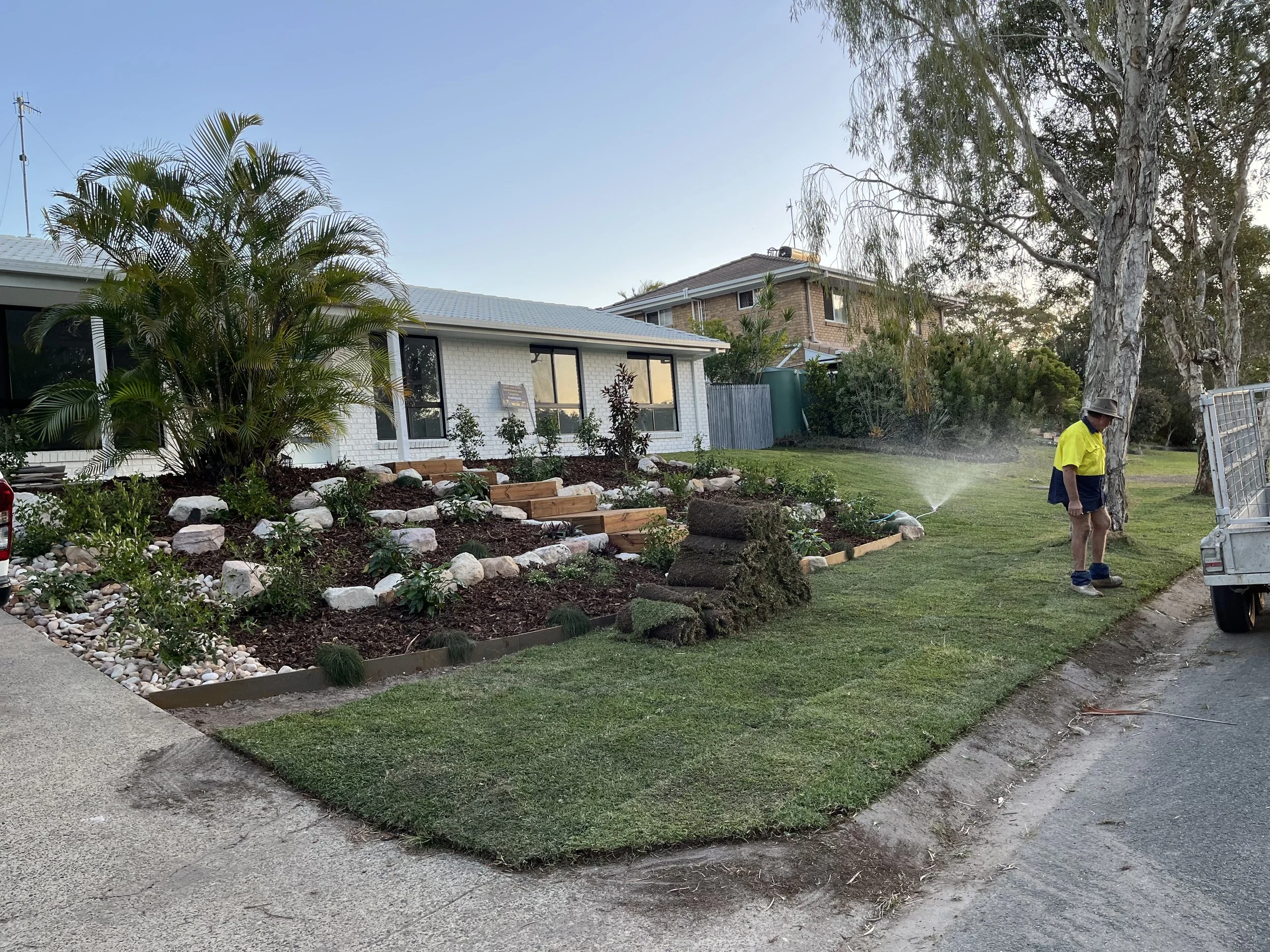 A backyard with a landscaped garden bed filled with plants and white rocks, next to a lawn and a house with large windows. A person wearing a yellow shirt, hat, and shorts is watering the garden.
