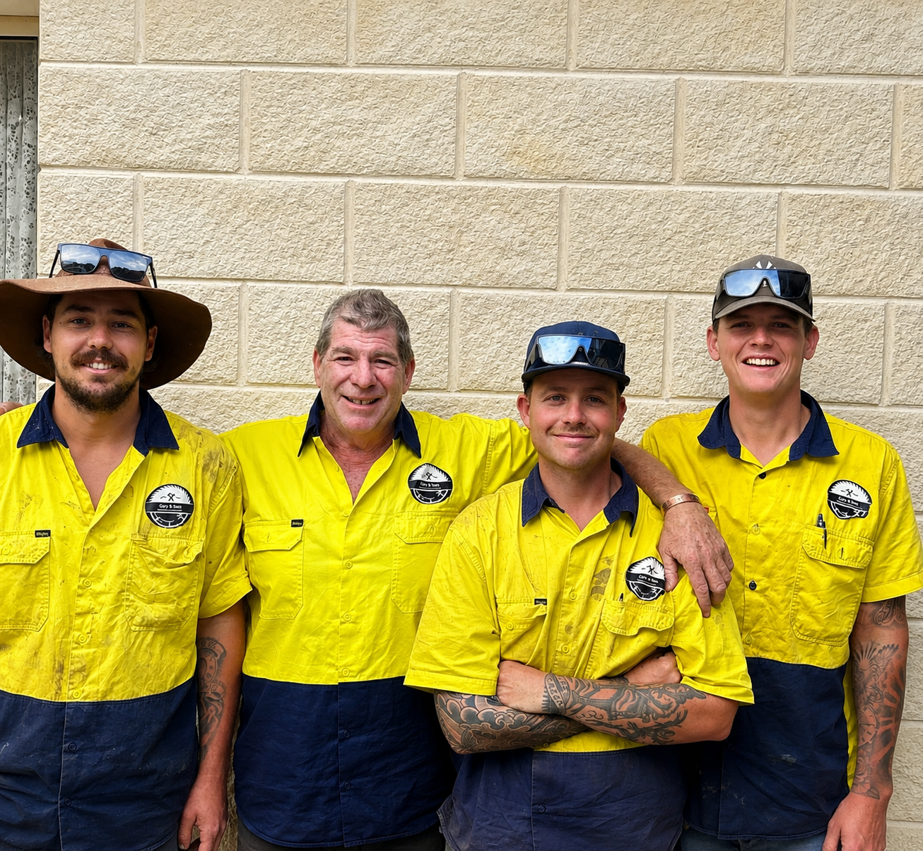 Four men wearing yellow and navy uniforms with logos, standing against a brick wall, smiling, with sunglasses on their heads.