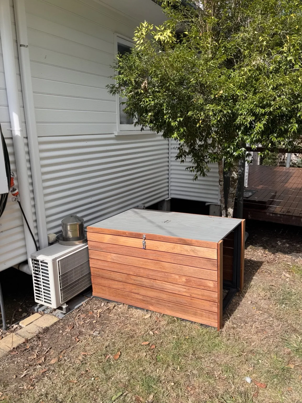 A small outdoor wooden storage box next to an air conditioning unit, with a tree and a deck in the background.