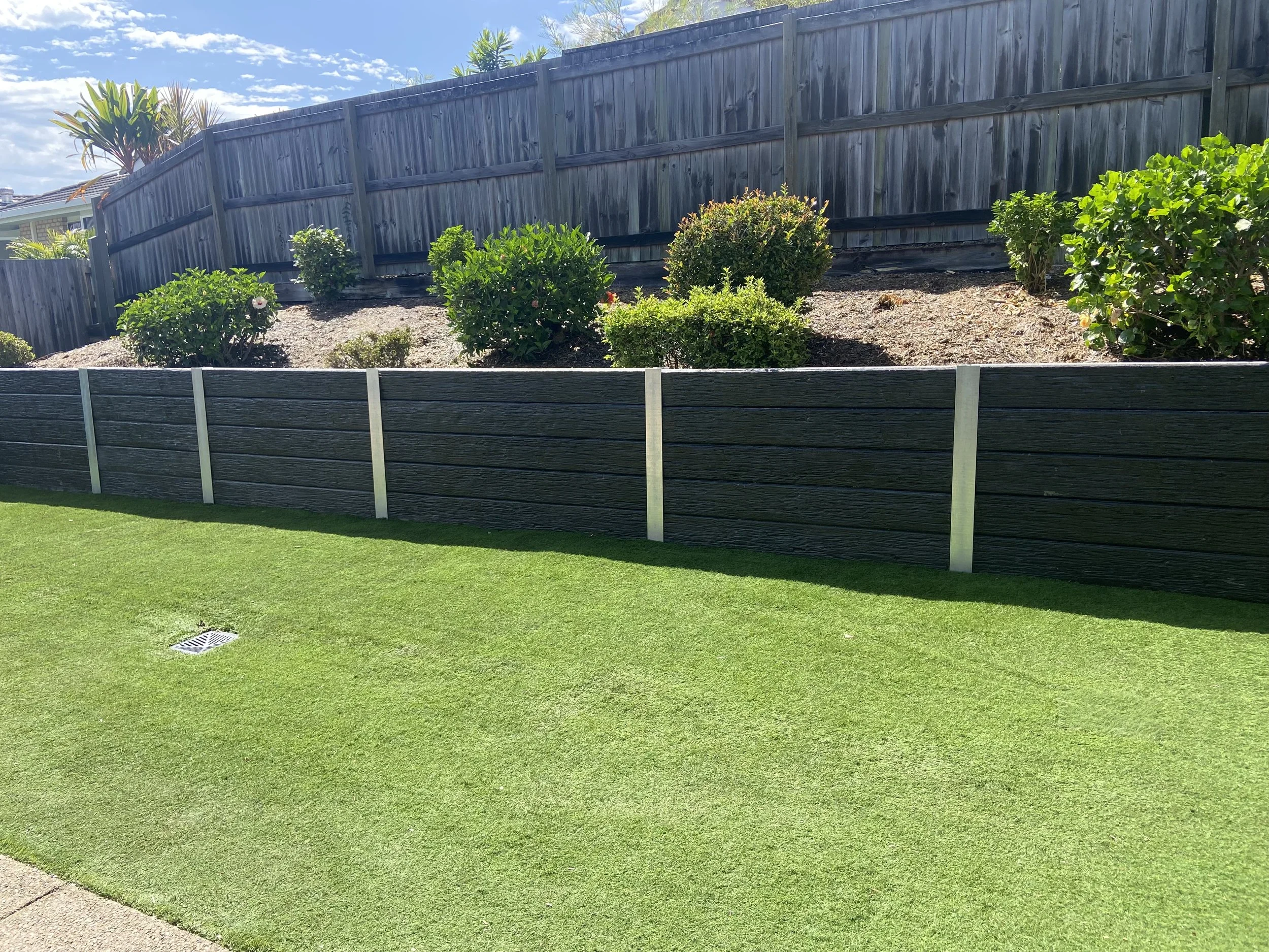 A backyard with a well-maintained green lawn, a black wooden fence, and a landscaped garden bed with shrubs and bushes, under a blue sky with scattered clouds.