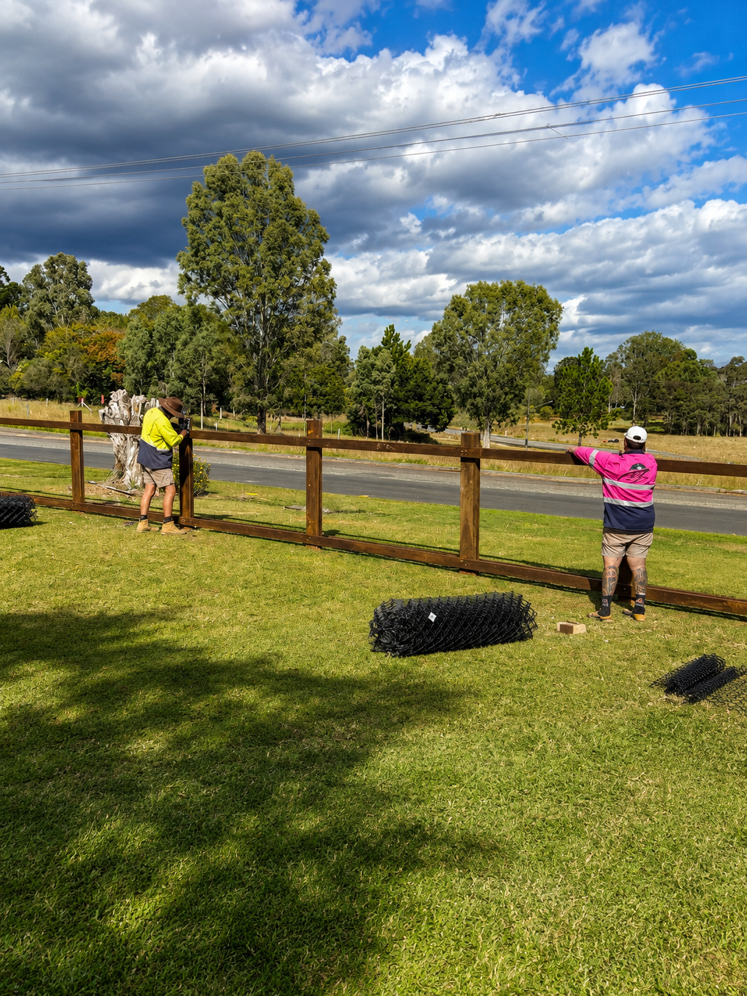 Two men installing a wooden fence along a grassy area beside a road, with trees and a cloudy sky in the background.