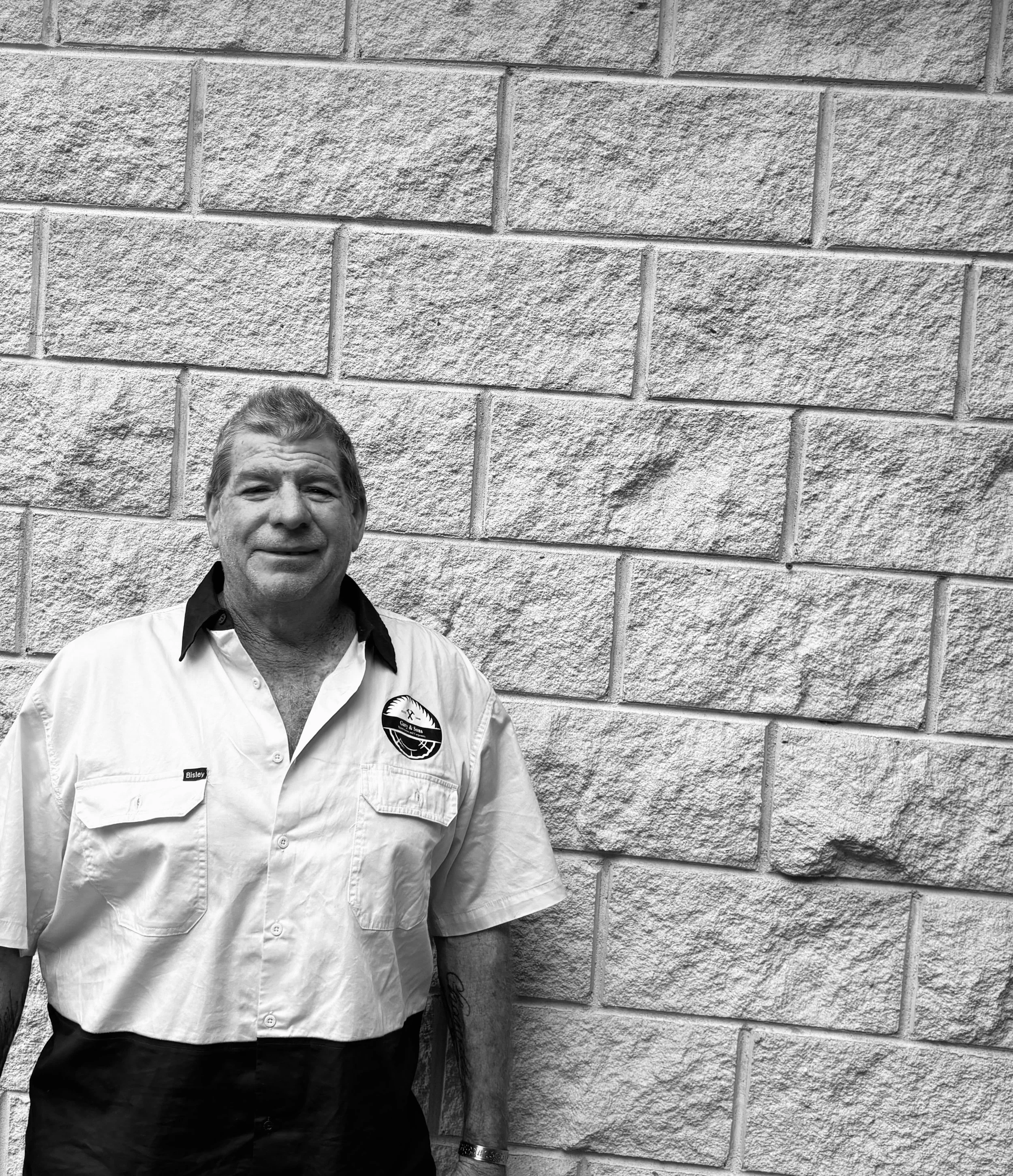 Black and white photo of a man with short hair and a smiling expression, standing against a brick wall. He is wearing a light-colored, short-sleeved shirt with a logo on the pocket.