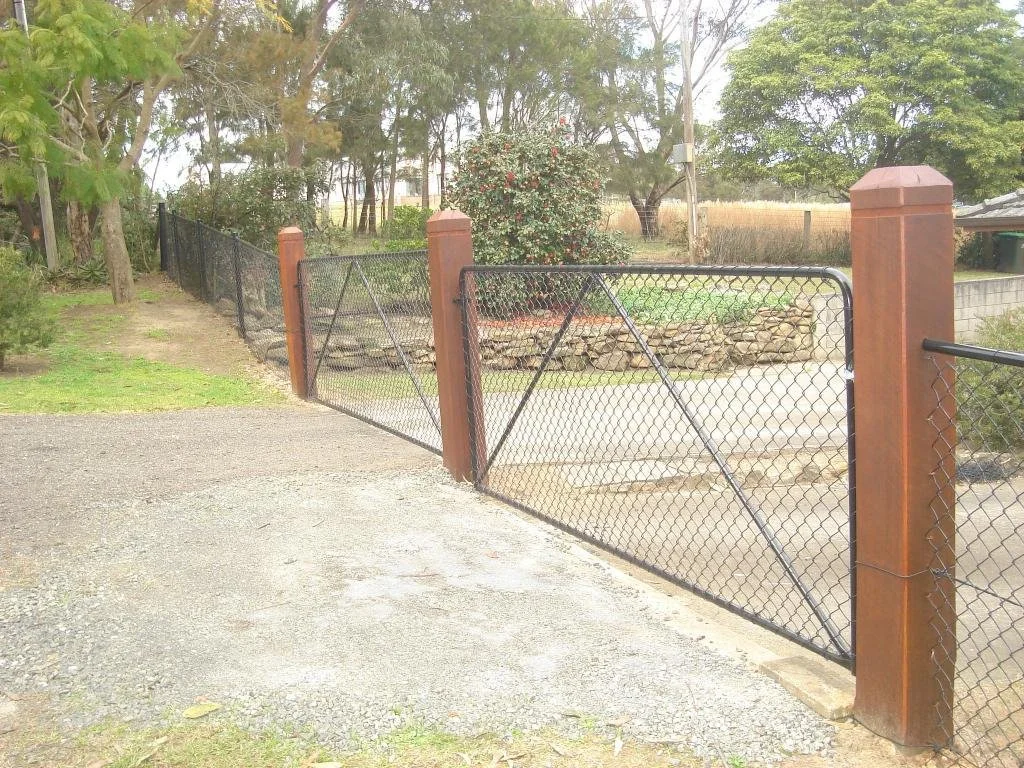 Black chain-link gate with brown metal posts leading into a yard with trees and bushes.