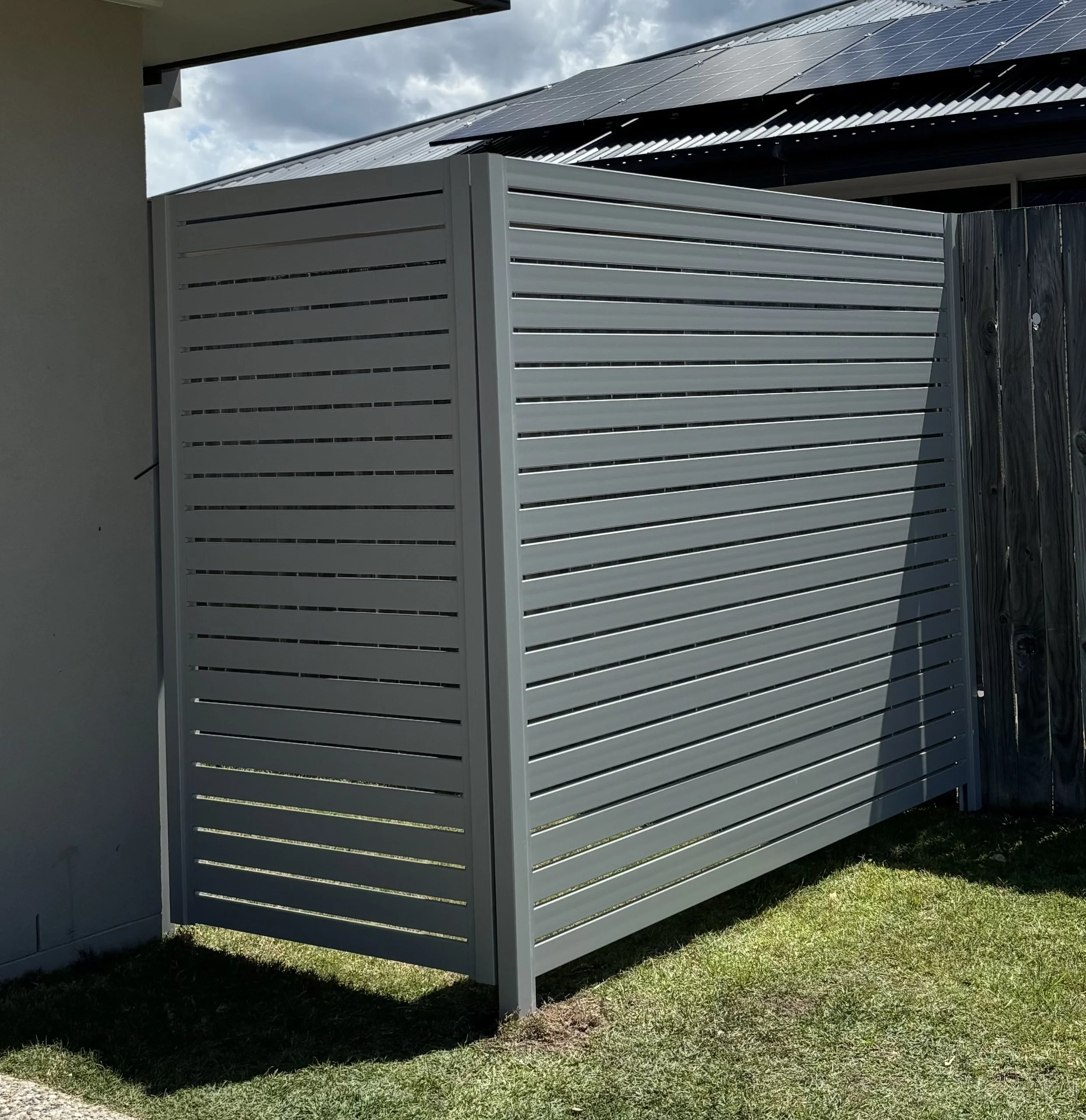 A white slatted outdoor privacy screen or fence in a backyard, with a metal roofed house in the background and partly cloudy sky.