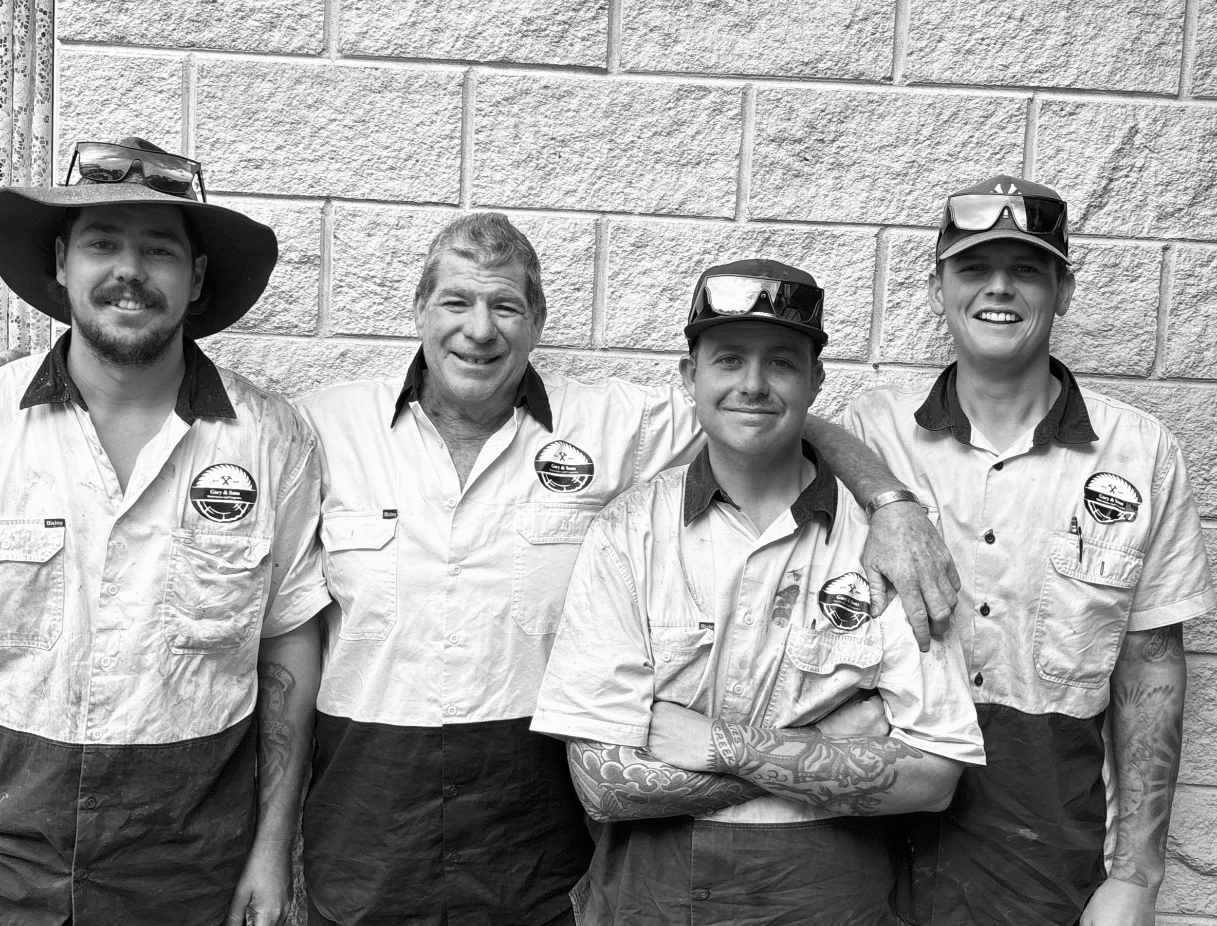 Four men standing against a brick wall, smiling, wearing casual shirts, some with sunglasses on their heads, one with a wide-brimmed hat, one with tattoos on their arms.