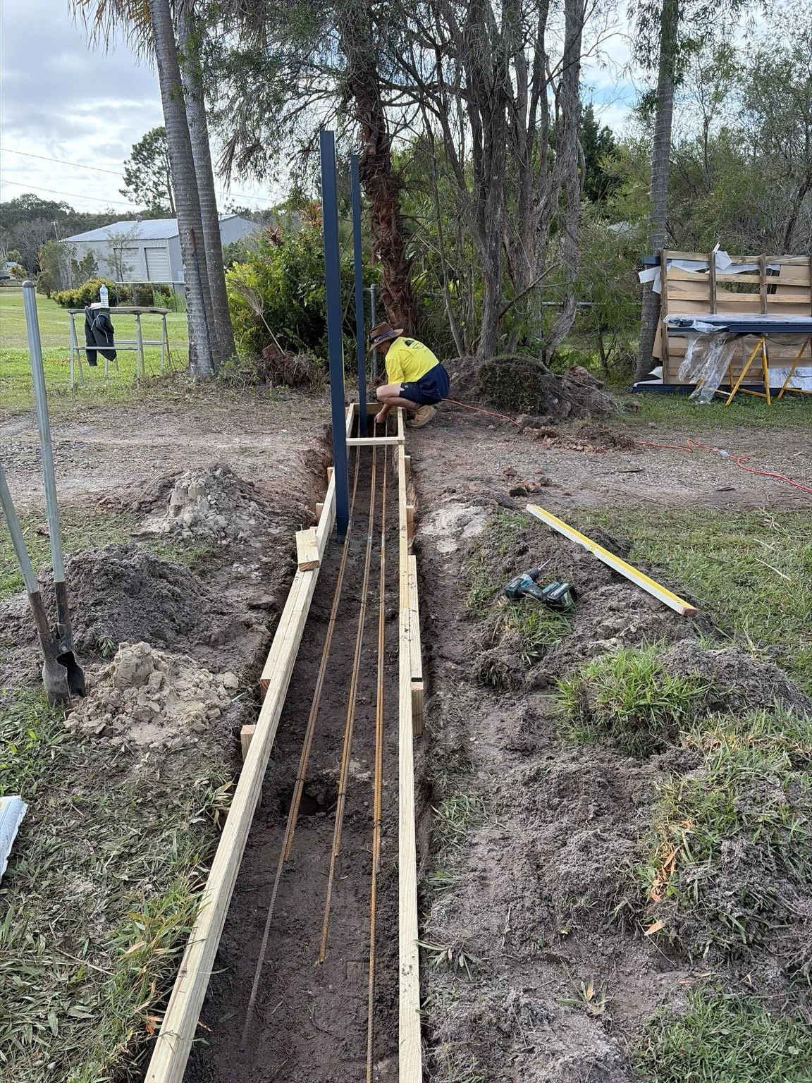 A person working on a construction project outdoors, installing metal rebar in a trench with wooden formwork, surrounded by trees and construction tools.