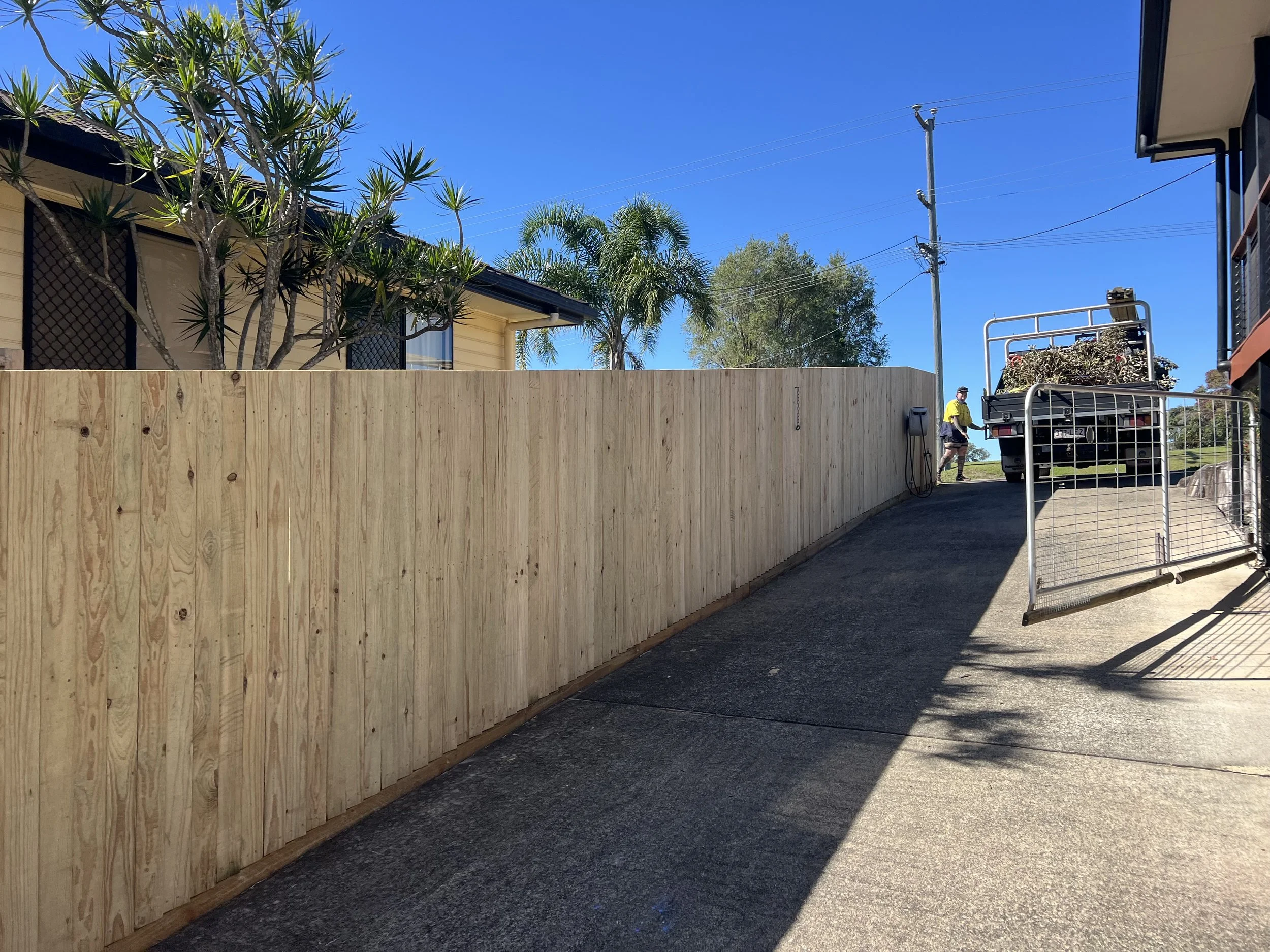 A wooden fence, with most of it visible, runs along the left side and bottom of the image. In the background, a house with beige siding and black window frames is partially visible. Several tall trees, including palm trees, are behind the house. A clear blue sky is overhead. On the right side, there is a flatbed truck with plants loaded onto it, parked near a person wearing a yellow shirt and shorts, who is standing near some equipment.