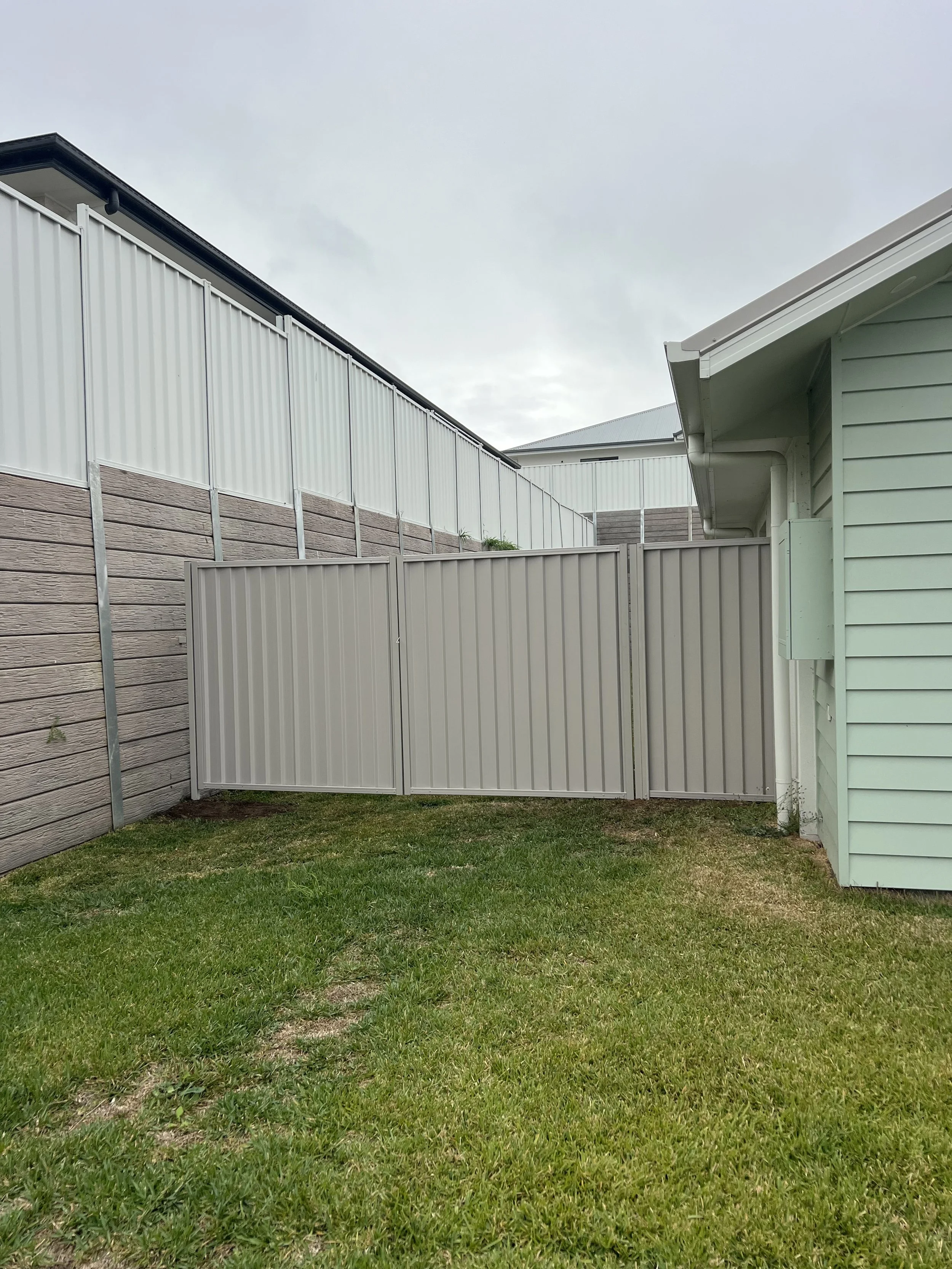 A backyard with a green grass lawn, a wooden fence on the left, a white metal fence on the right, and a corner of a light green house with siding. Overcast sky in the background.