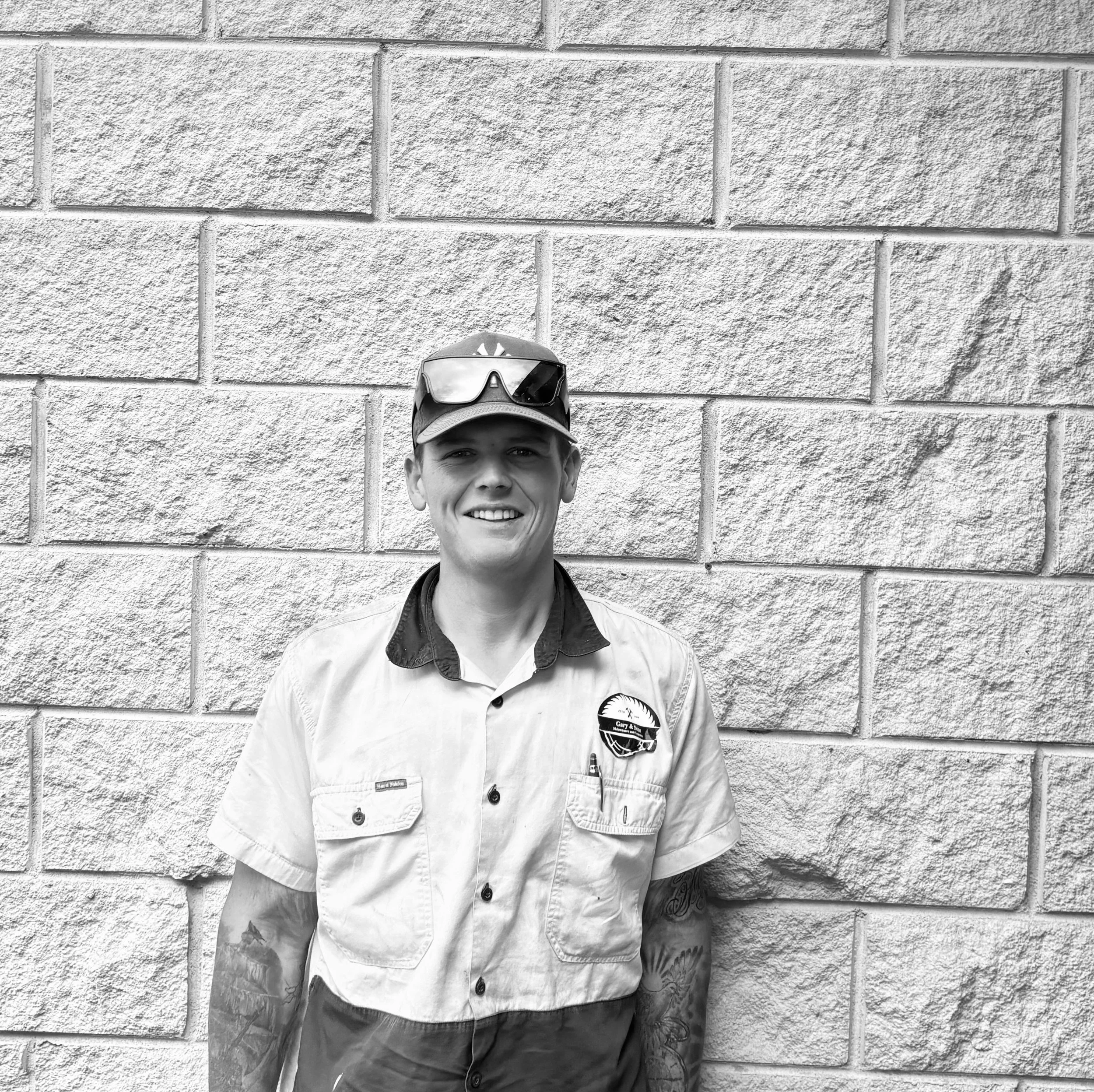 Portrait of a young man wearing a baseball cap and sunglasses, standing in front of a brick wall.