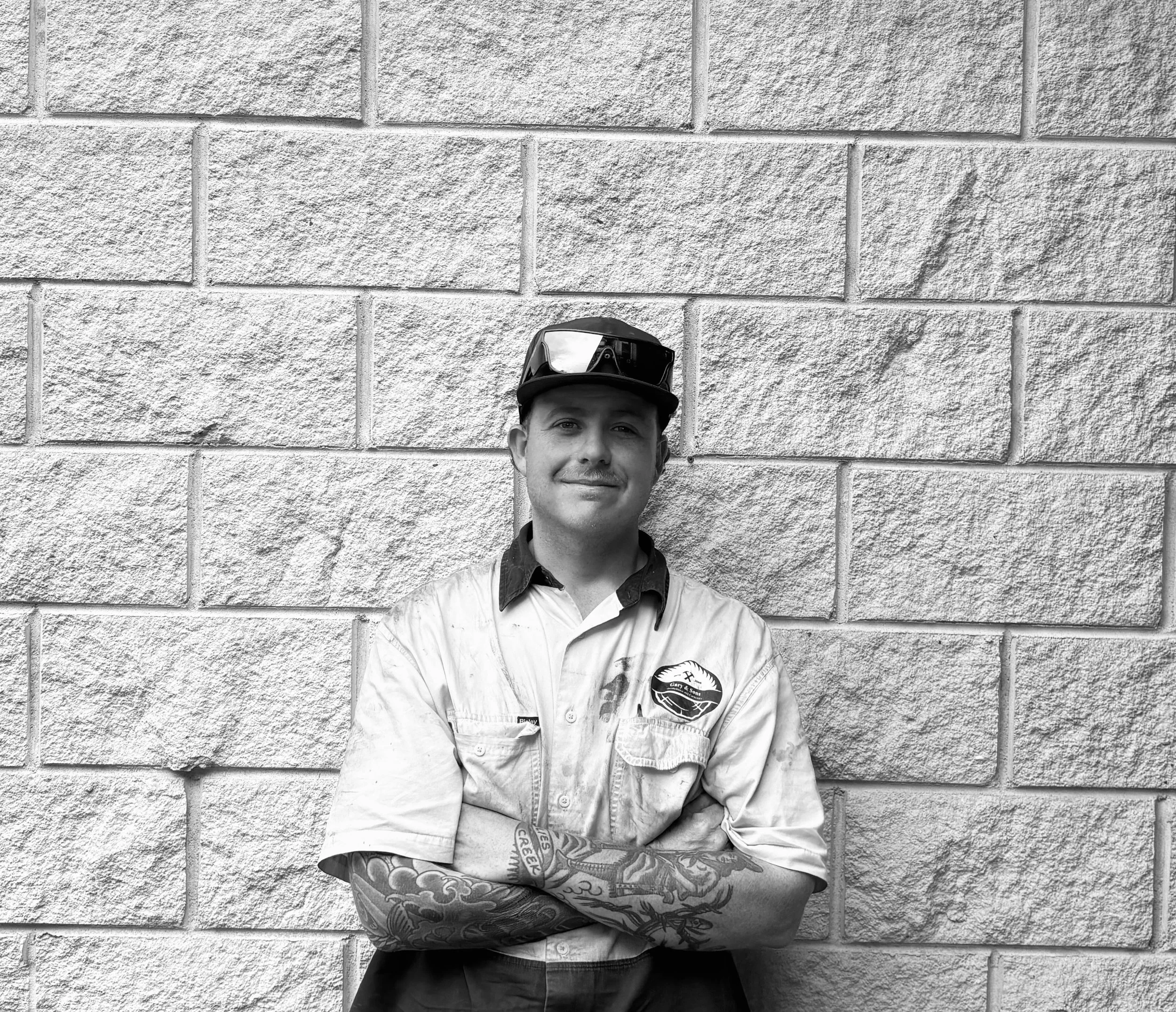 Black and white photo of a man wearing a baseball cap with sunglasses on top, standing against a brick wall. The man has tattoos on his arms and is smiling.