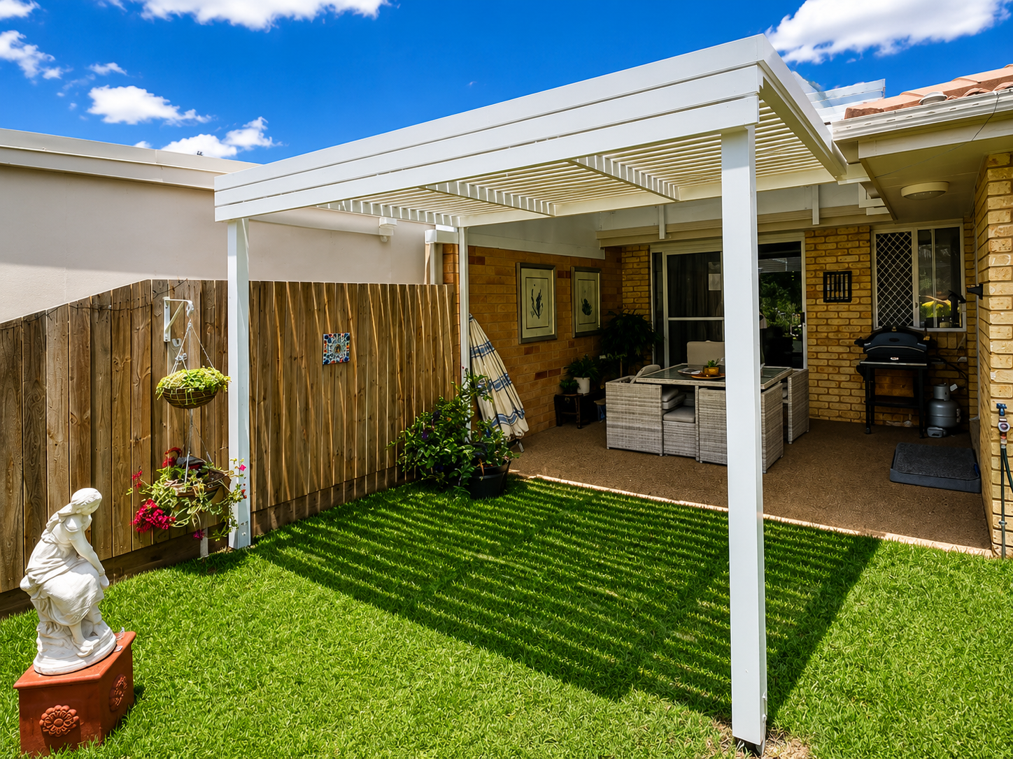 Backyard patio with a white pergola, green grass, potted plants, a statue, and a barbecue grill, with a brick house wall in the background.