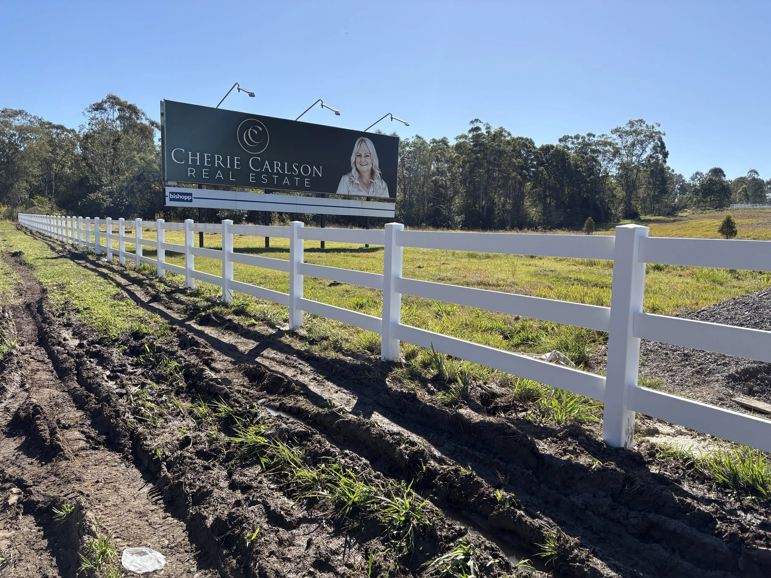 Billboard advertising Cherie Carlson Real Estate next to a white fence, with green trees and clear blue sky in the background.