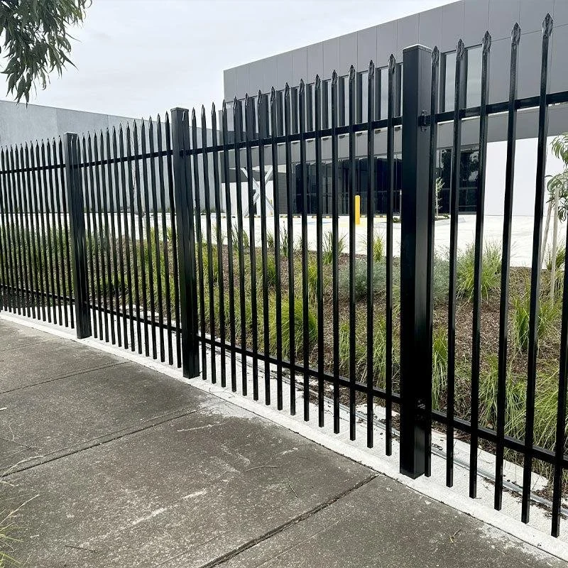 Black metal fence along sidewalk with building behind it and greenery inside the fenced area.