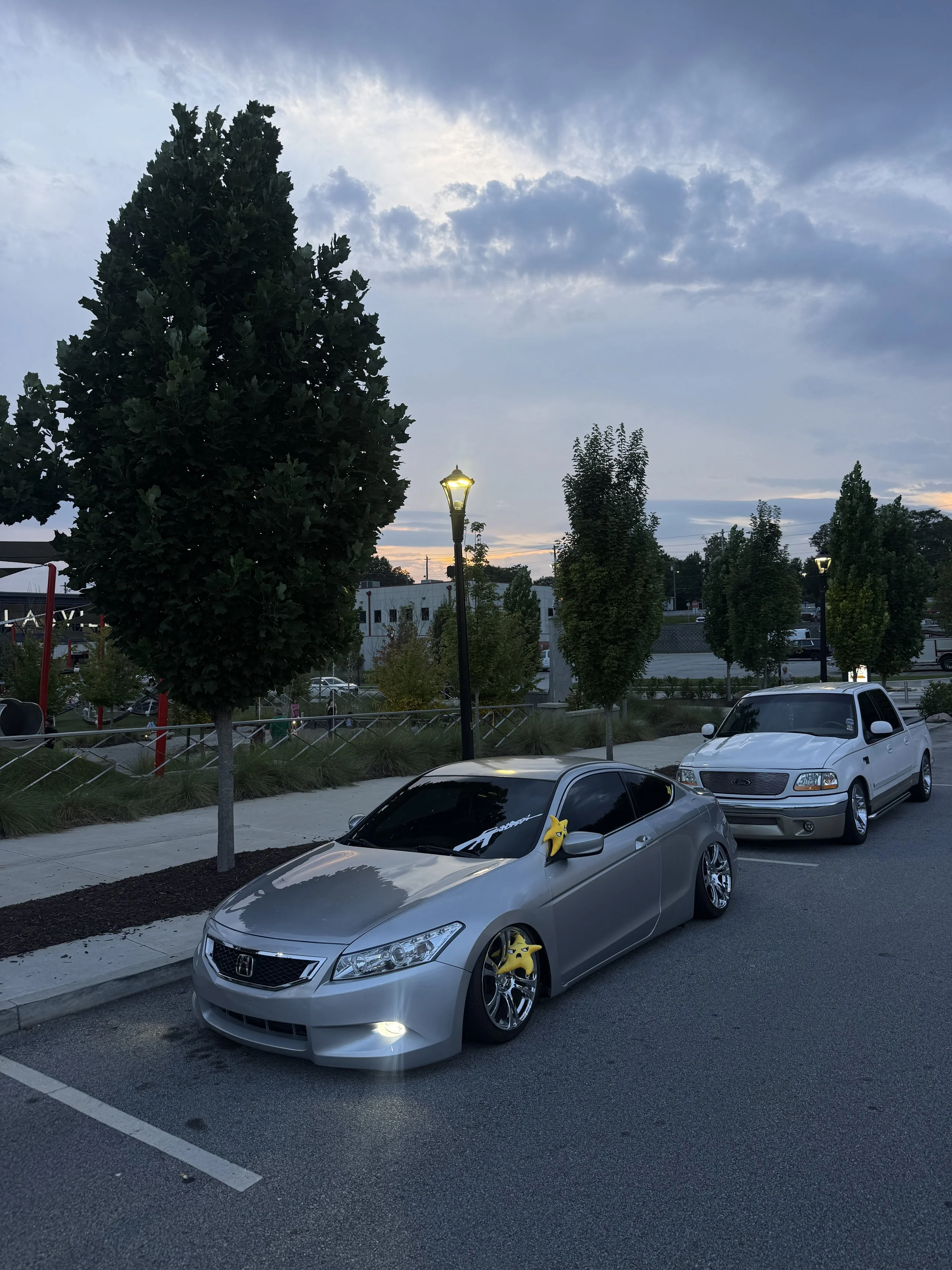 A parking lot with a silver car in the foreground, decorated with yellow star-shaped balloons on the side mirrors. Other cars are parked nearby, and there are trees and street lamps along the parking area. The sky is cloudy with some light breaking through as evening approaches.