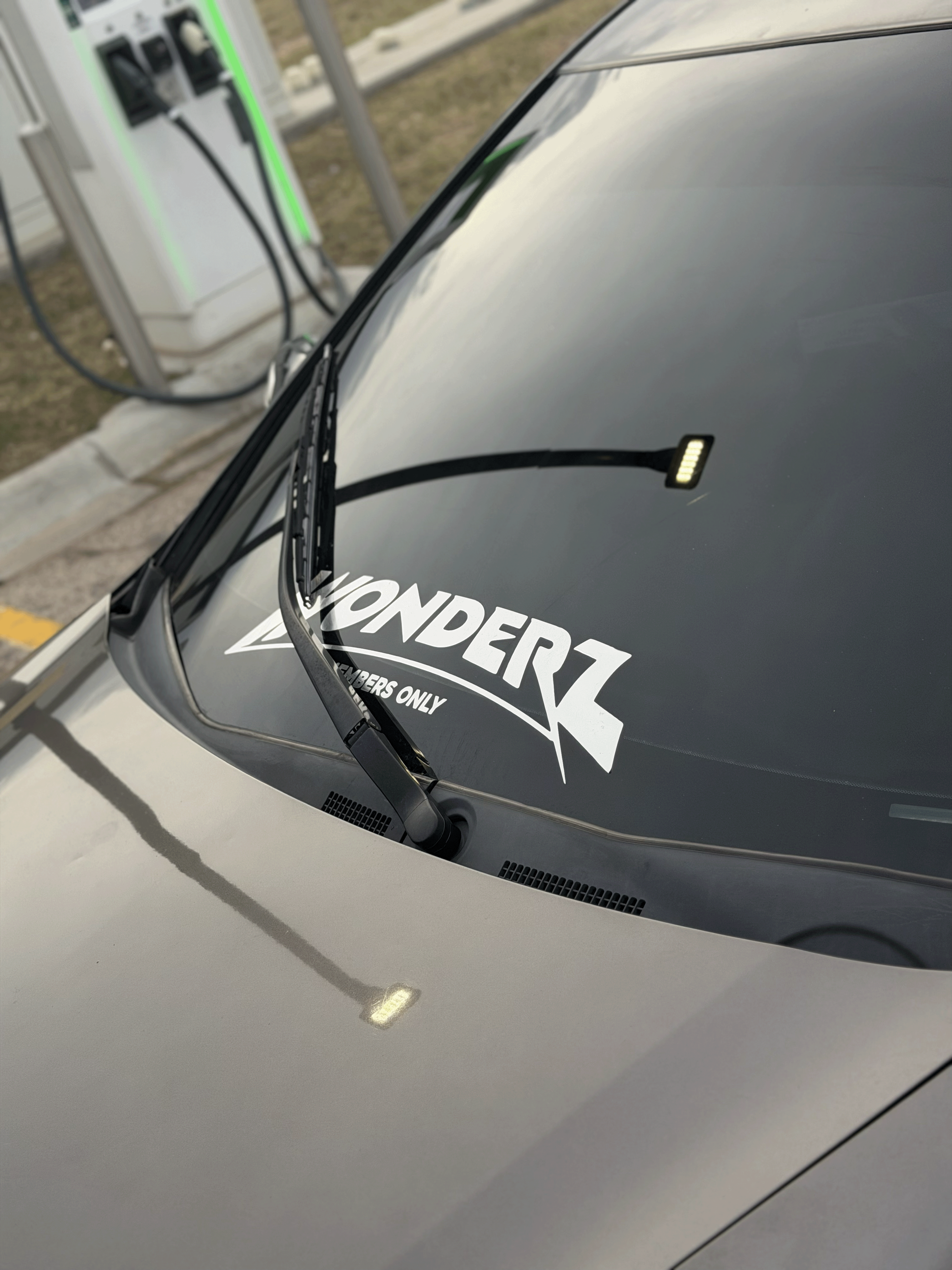 Close-up of a silver electric vehicle with a black windshield sticker reading 'WONDERZ' and a reflection of a charger on the hood, outdoors