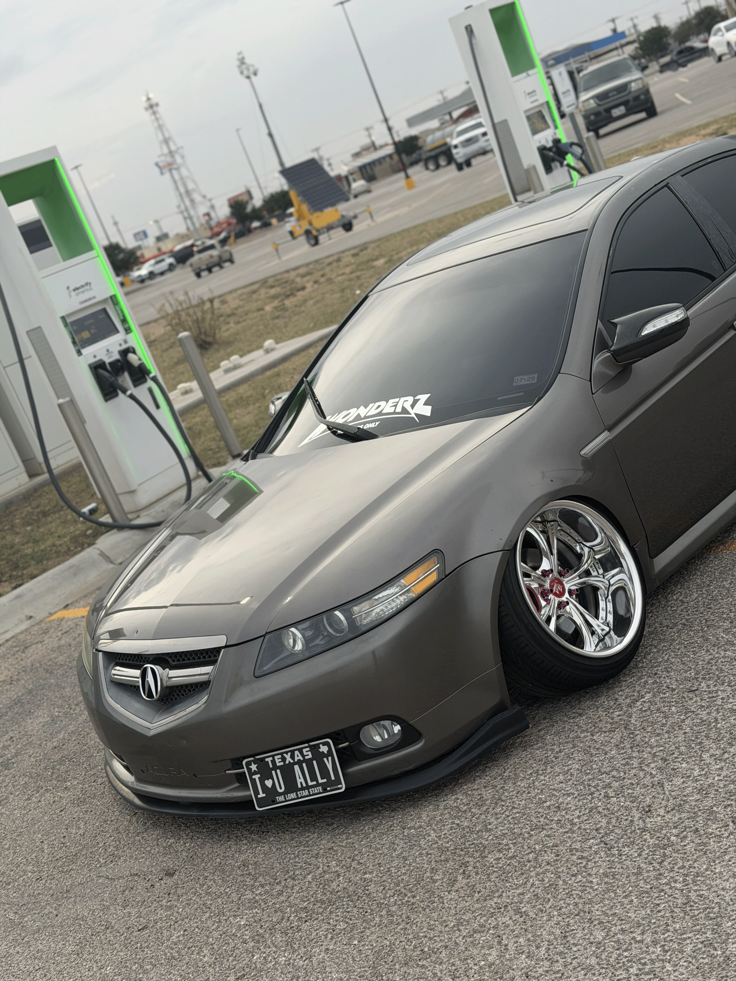 A silver Acura car with custom wheels parked at an electric vehicle charging station. The car has a windshield decal that says 'WONDERZ'.