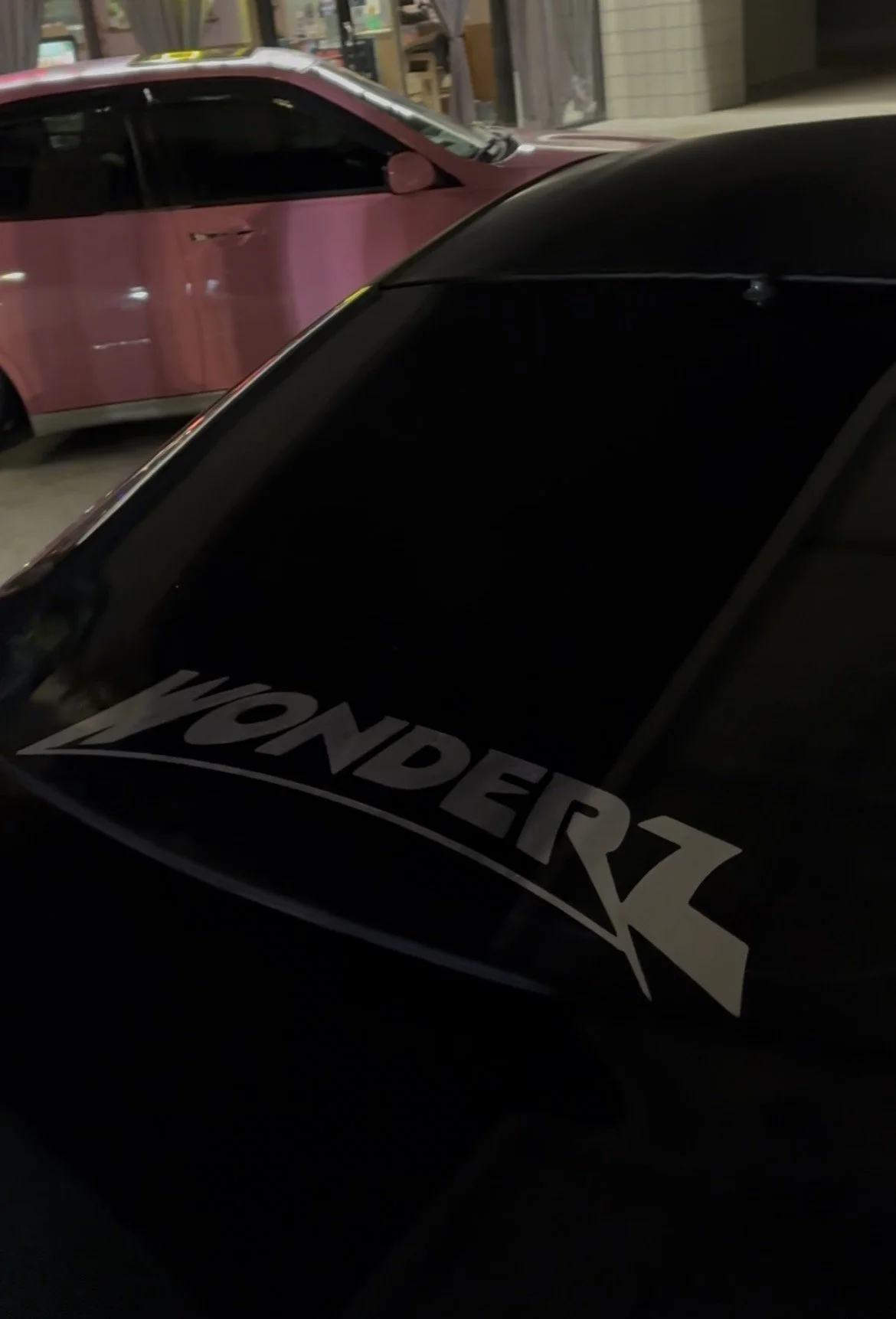 A black vehicle with a sticker that says 'YANDERZ' on the hood, parked indoors with a pink car and storefront in the background.