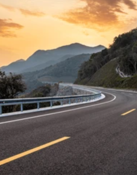 A winding mountain road at sunset with guardrails and rolling hills in the background.