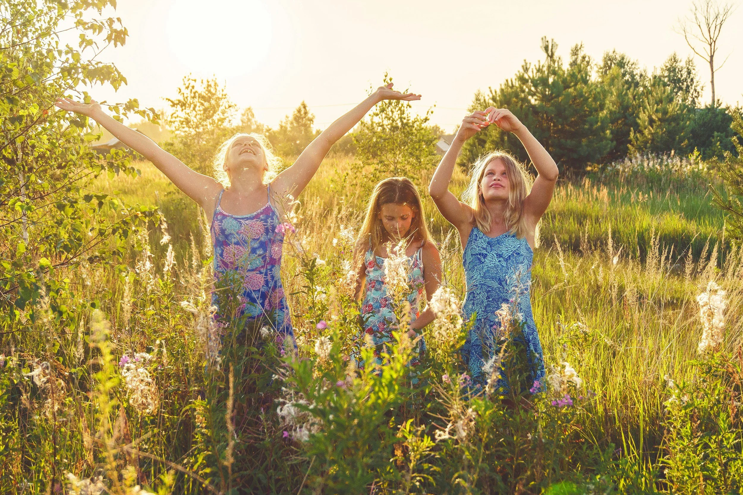 Three young girls in colorful dresses joyfully playing and dancing in a grassy field with wildflowers during a sunny late afternoon or early evening.