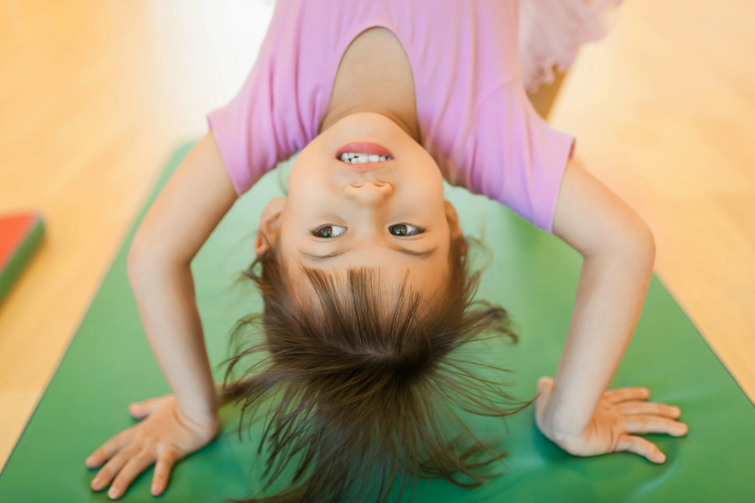 A young girl with brown hair and brown eyes lying on her stomach on a multicolored yoga mat, smiling upside down at the camera, with her hands positioned in a child's pose.