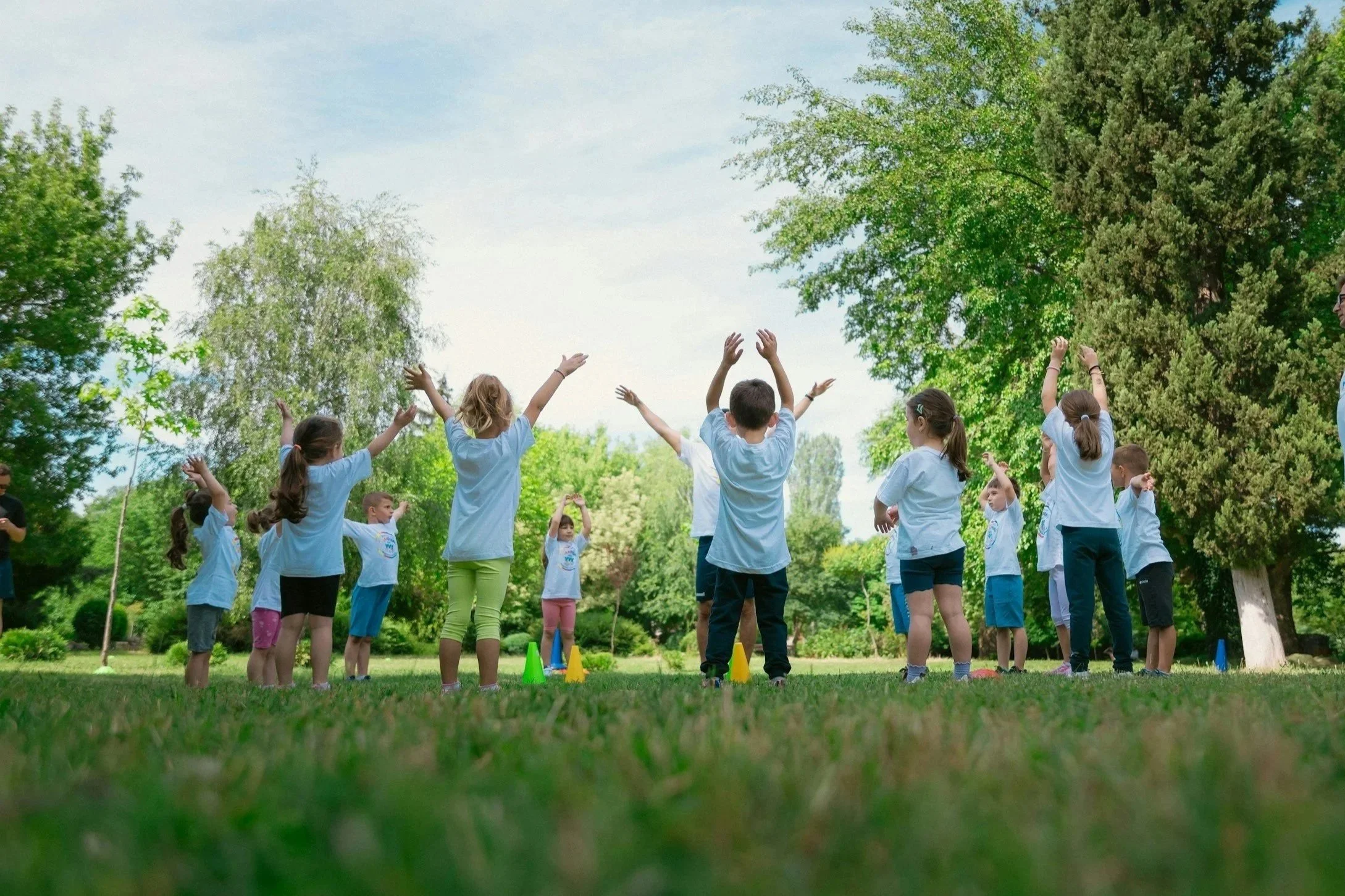 Children participating in outdoor activities in a park with lush green trees and a clear sky.