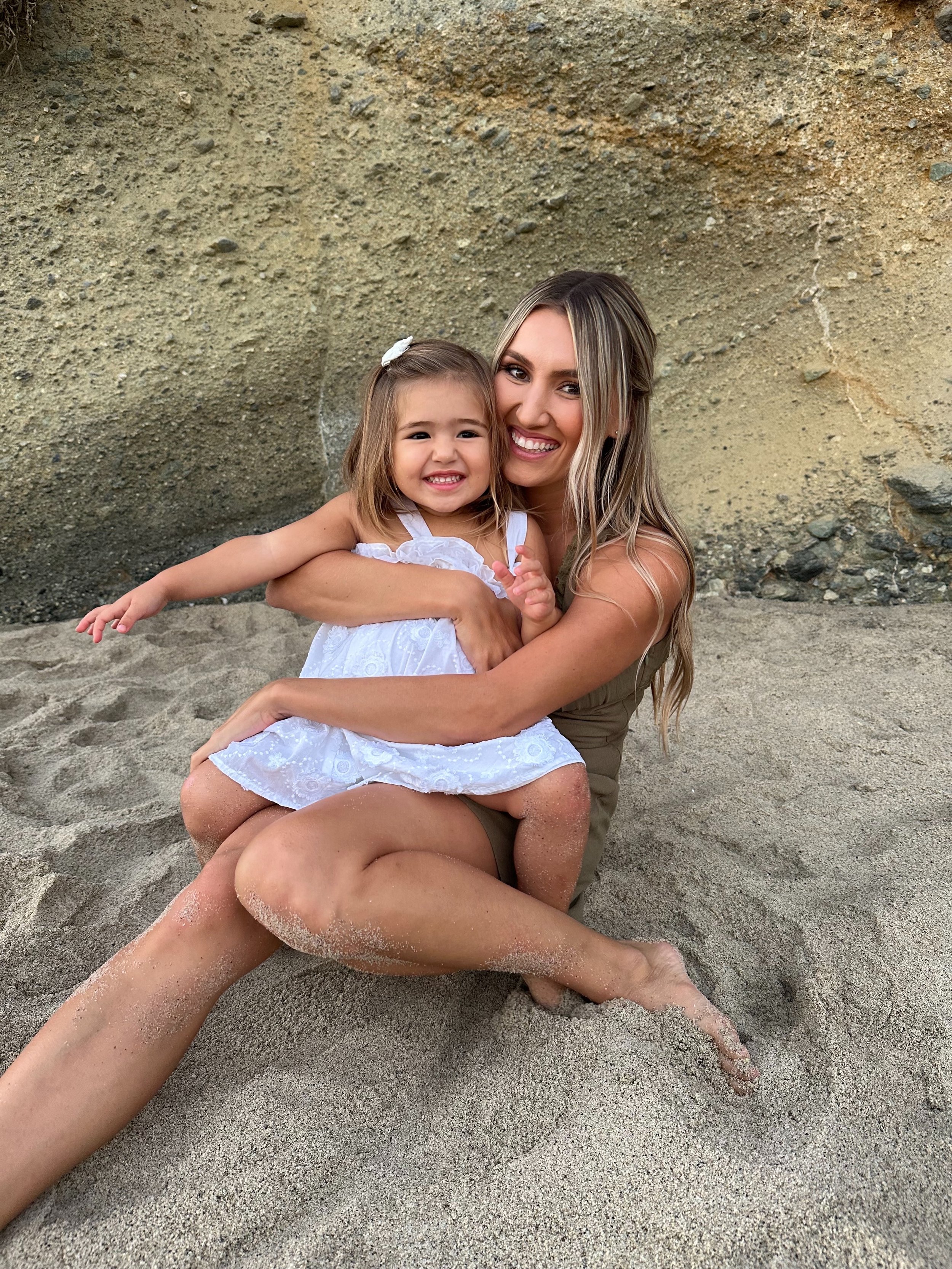 A woman and a young girl sitting on the sandy beach, smiling and embracing each other against a large sandy rock wall.